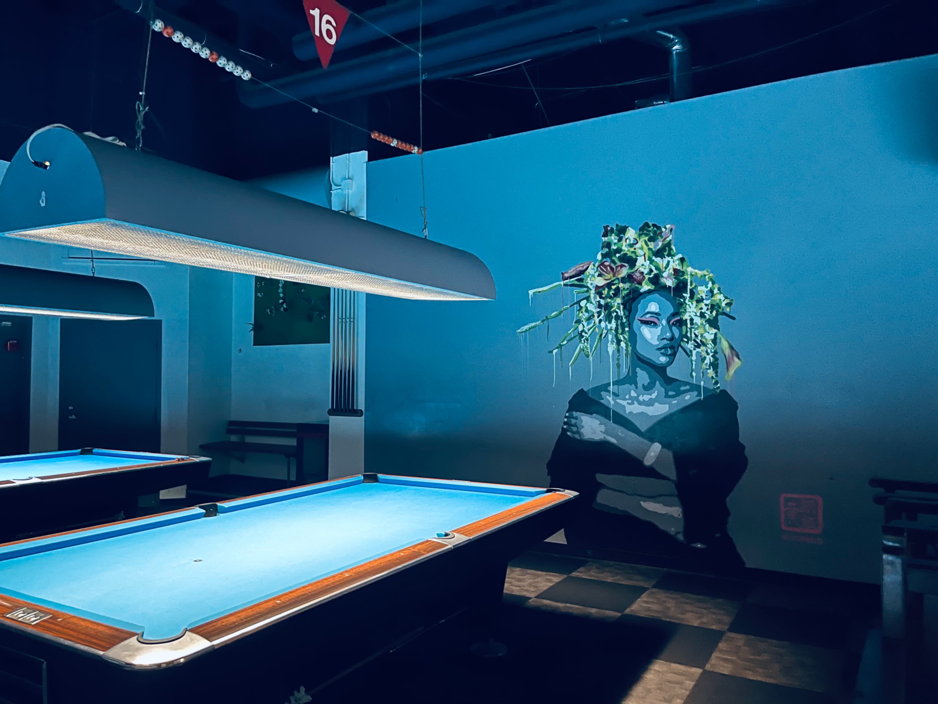 A pool hall featuring a vibrant mural of a woman with greenery on her head next to a blue-felted pool table.