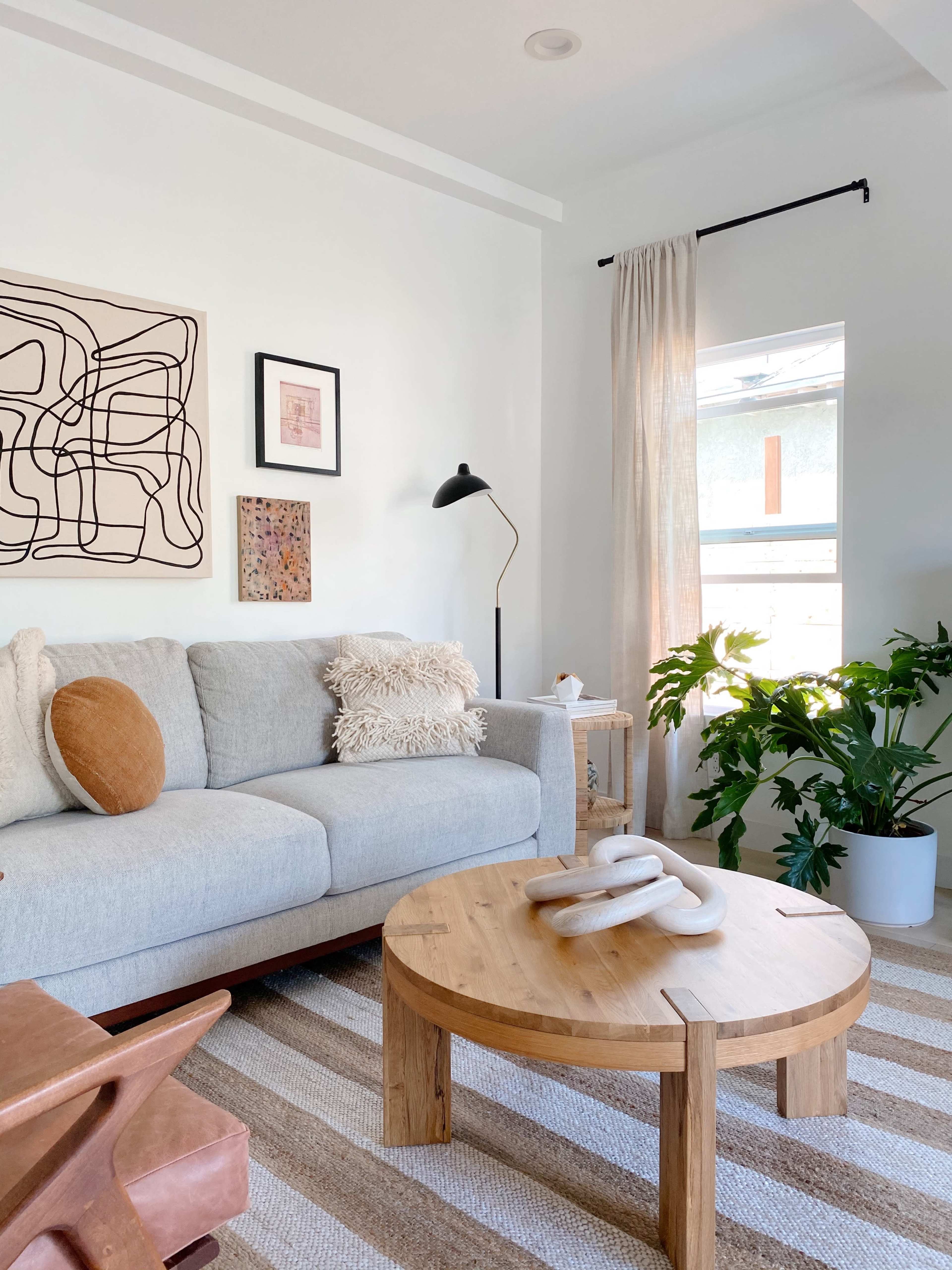 A modern living room features a light gray sofa, a round wooden coffee table, and a large potted plant beside a window with sheer curtains.