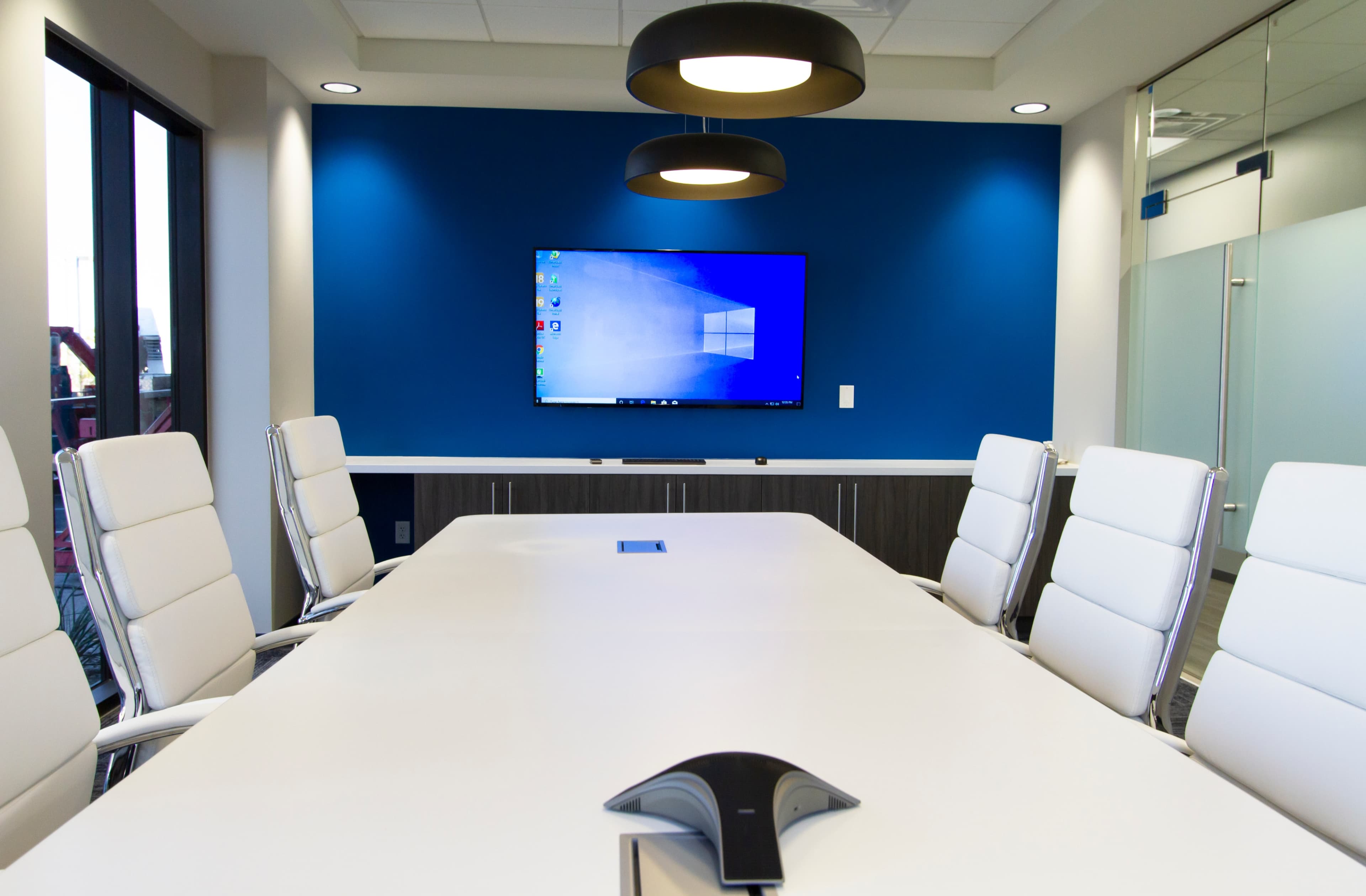 A bright conference room features a long white table, white leather chairs, and a large wall-mounted screen against a blue backdrop.