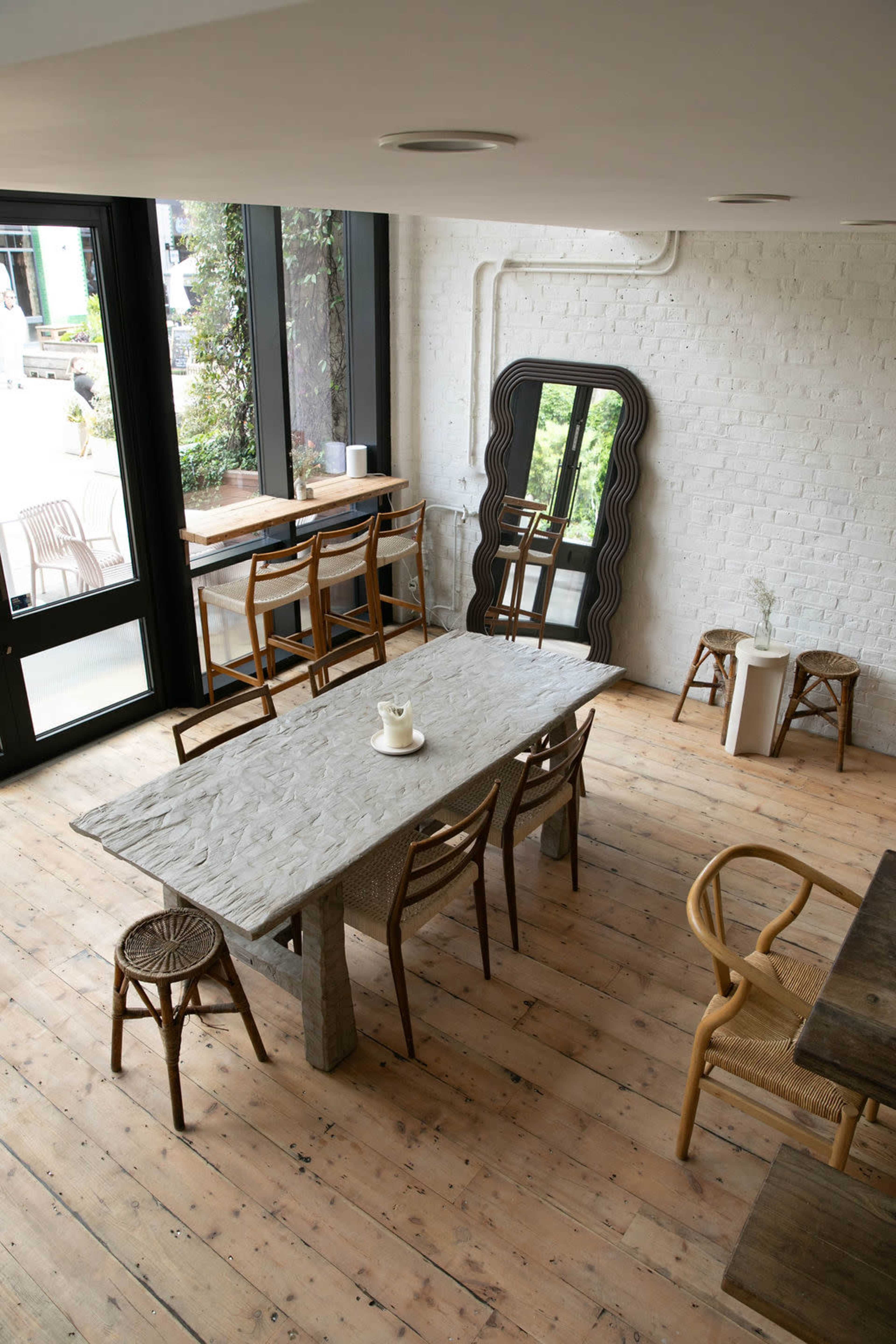 The image shows a modern dining area with a large stone table surrounded by wicker chairs, complemented by a full-length mirror and large windows.