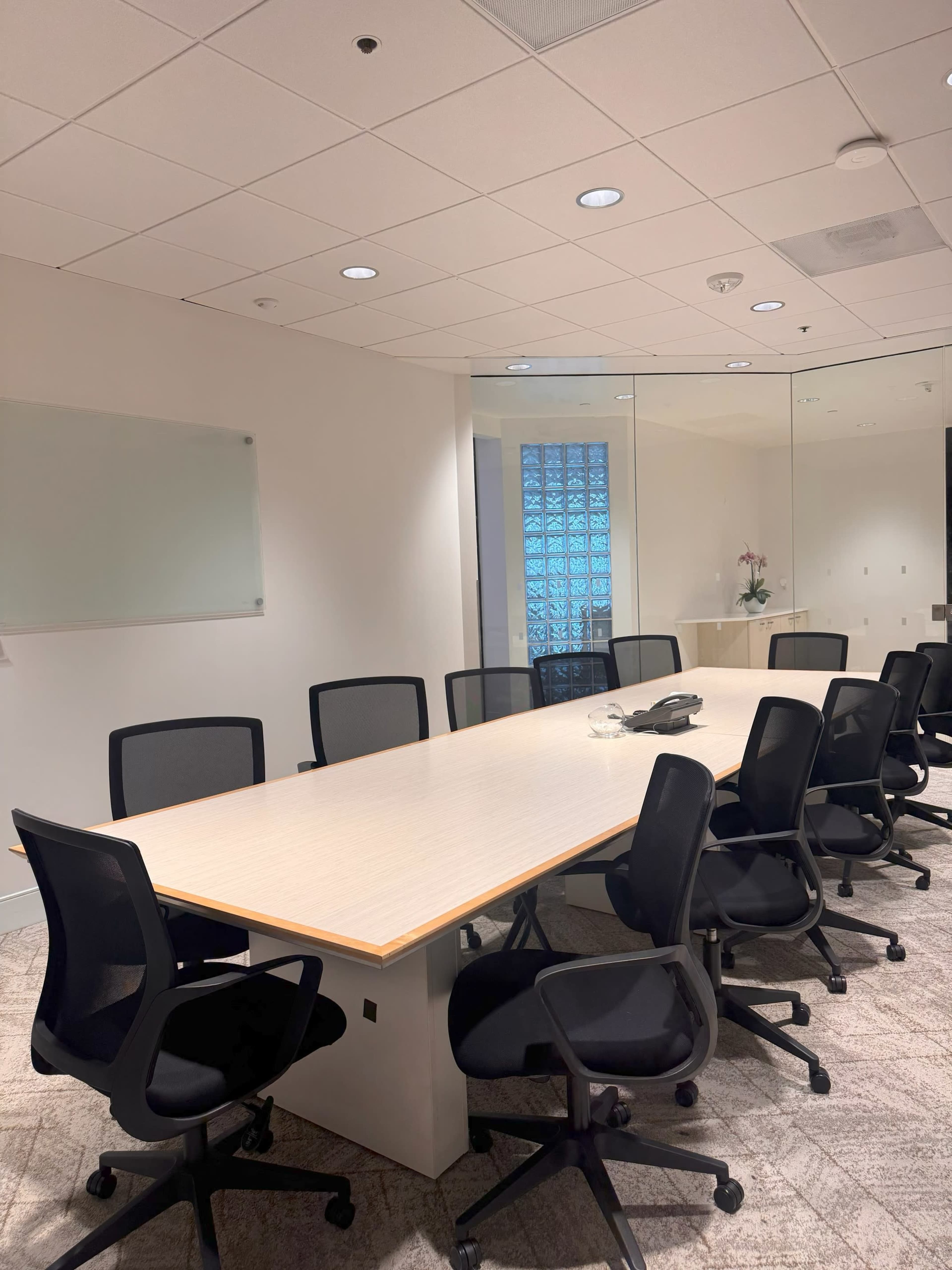 A modern conference room features a large, rectangular table surrounded by black office chairs, with a glass wall and a whiteboard visible in the background.