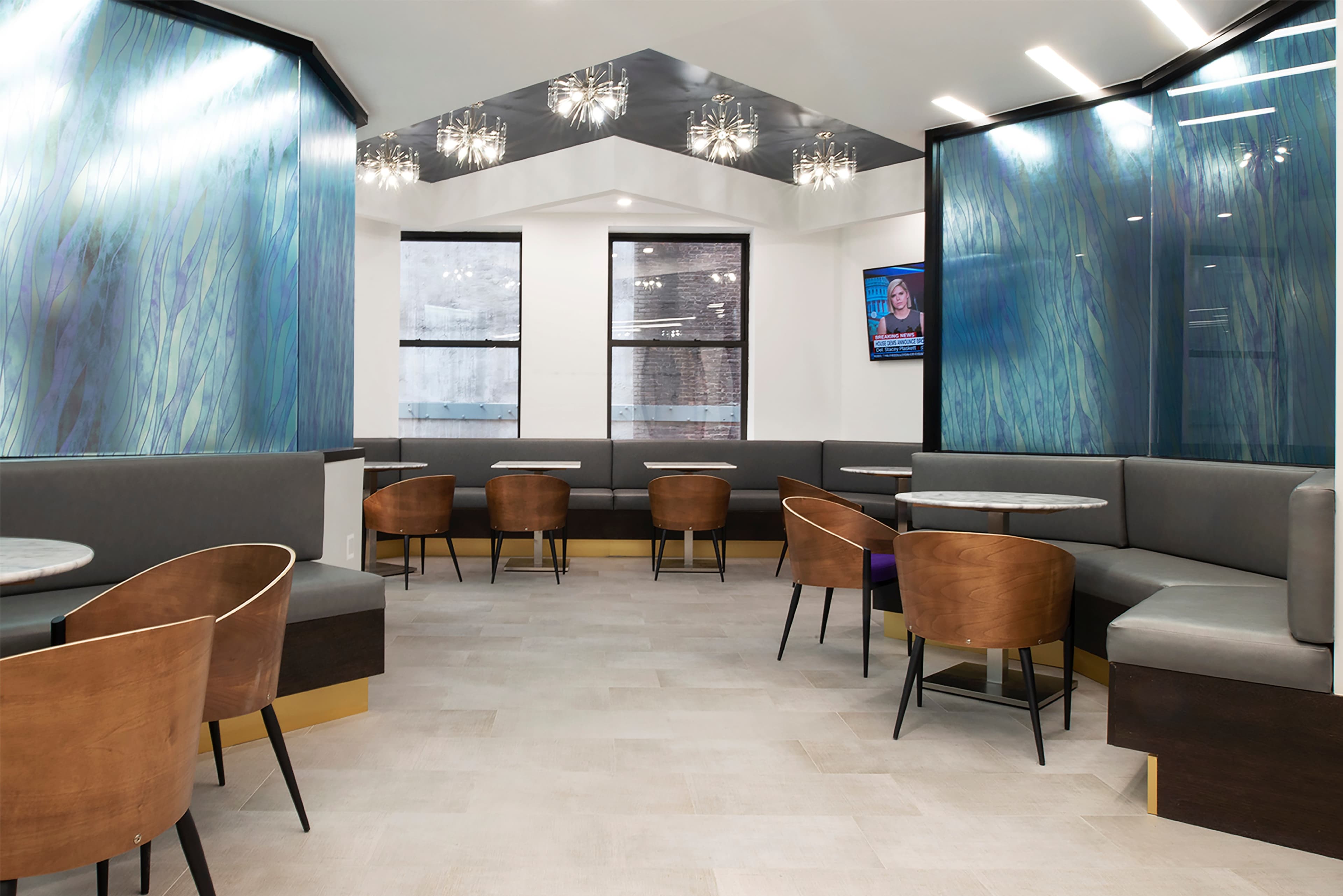 The image shows a modern, brightly lit lounge area with gray seating, wooden chairs, and large windows, featuring decorative light fixtures and a television on the wall.