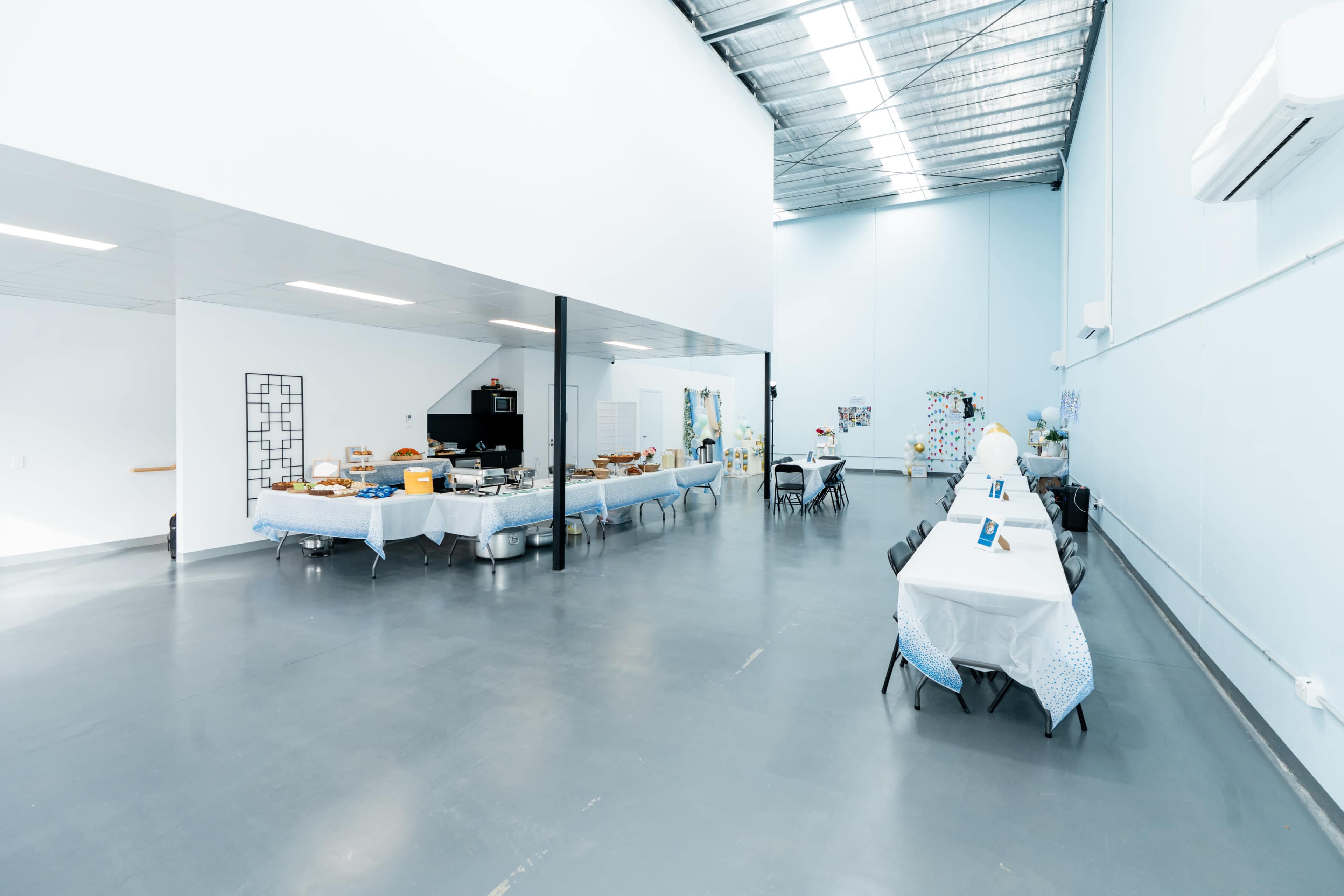 The image shows a spacious, well-lit event space with tables covered in white tablecloths arranged alongside a buffet setup.