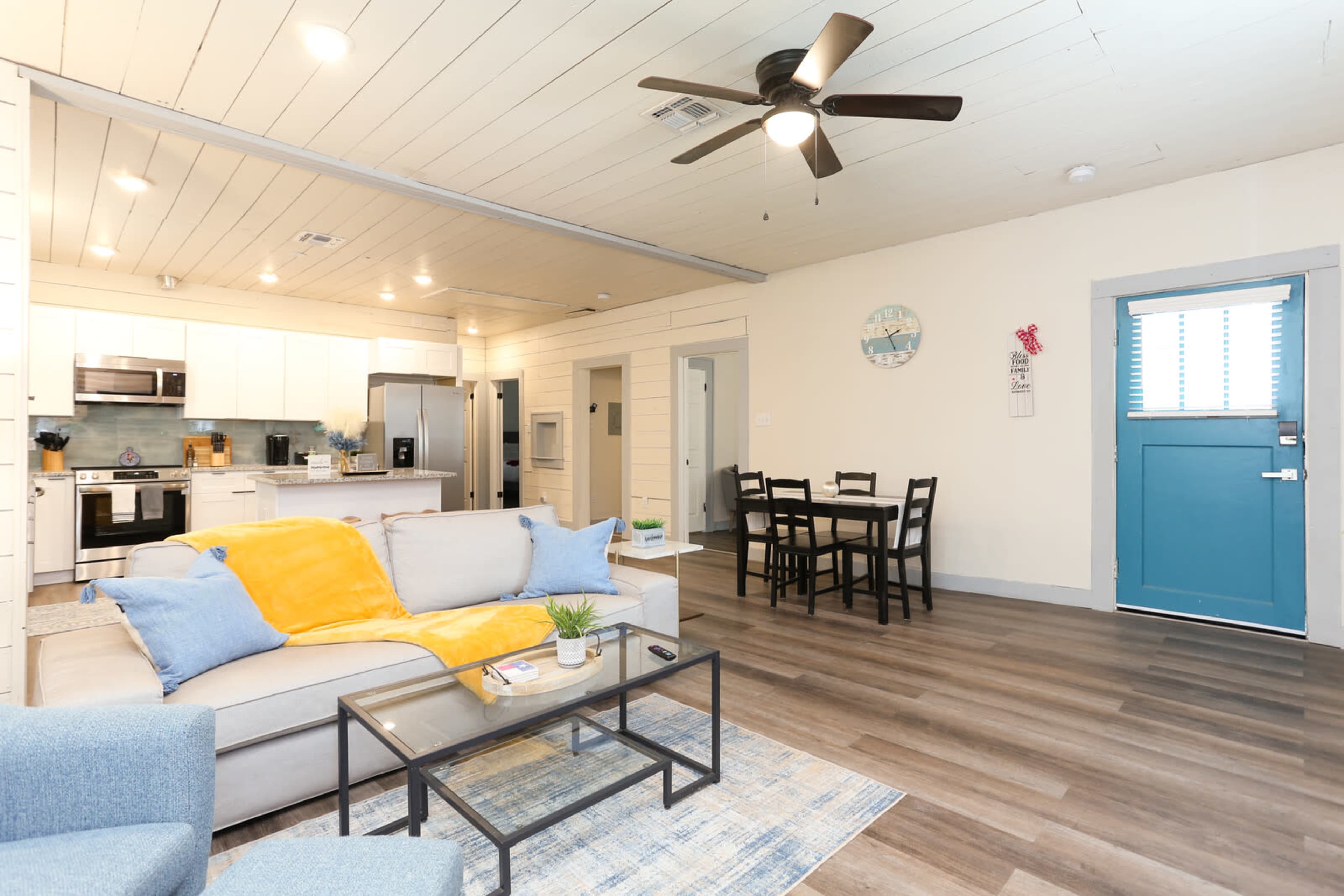 The image shows a modern living space featuring a light-colored sofa with a blue throw, a coffee table, a dining area with four chairs, and a blue door leading outside.