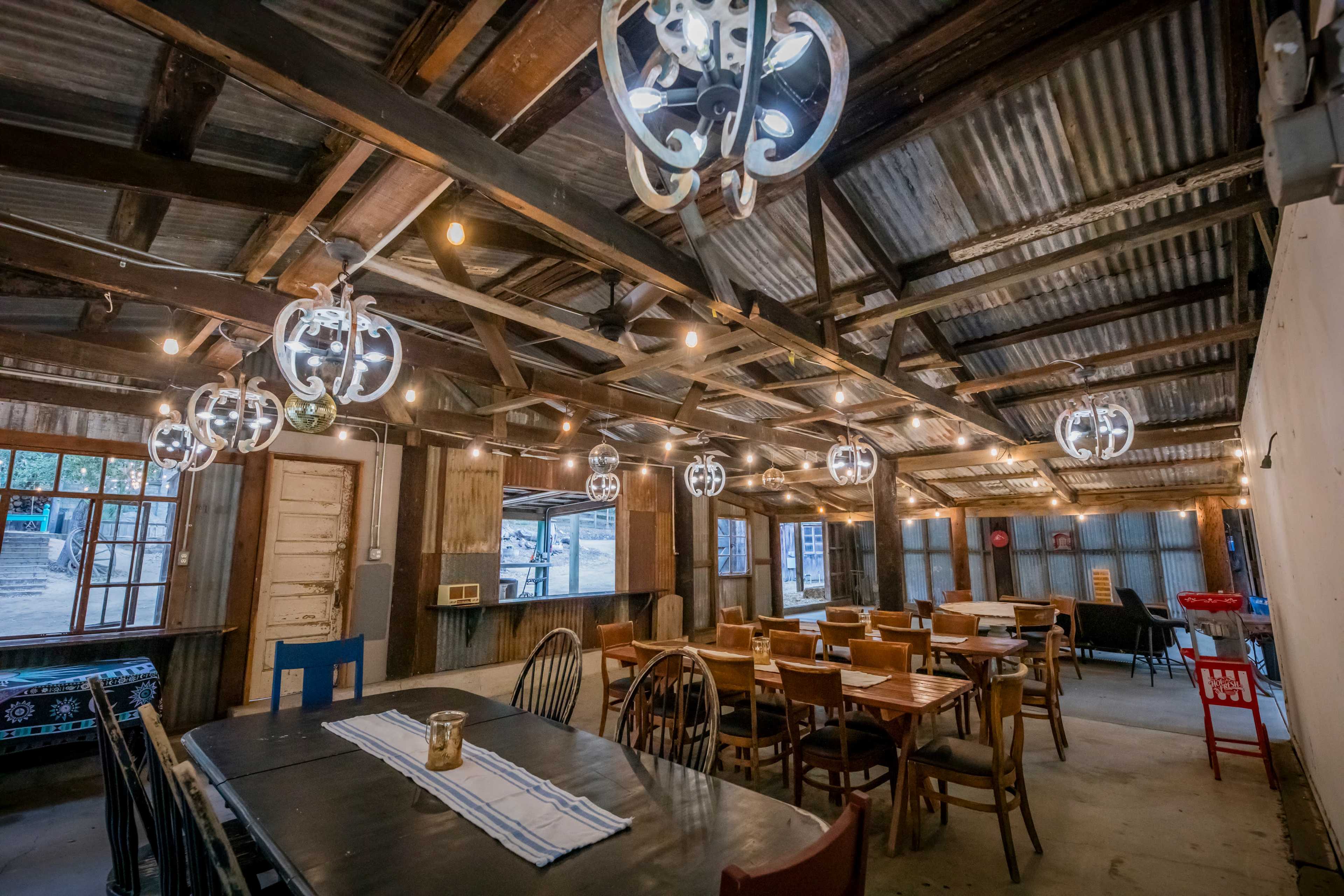 The image shows the interior of a rustic dining area with wooden tables and chairs, industrial-style light fixtures, and exposed beams.
