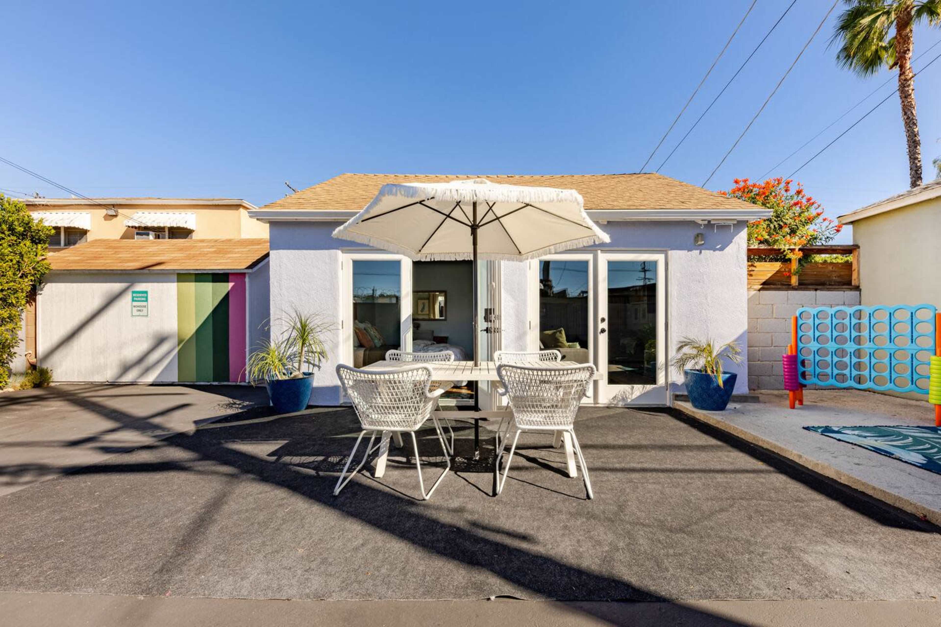A small, white house with large windows and a patio table under an umbrella is surrounded by a paved area and colorful landscaping.