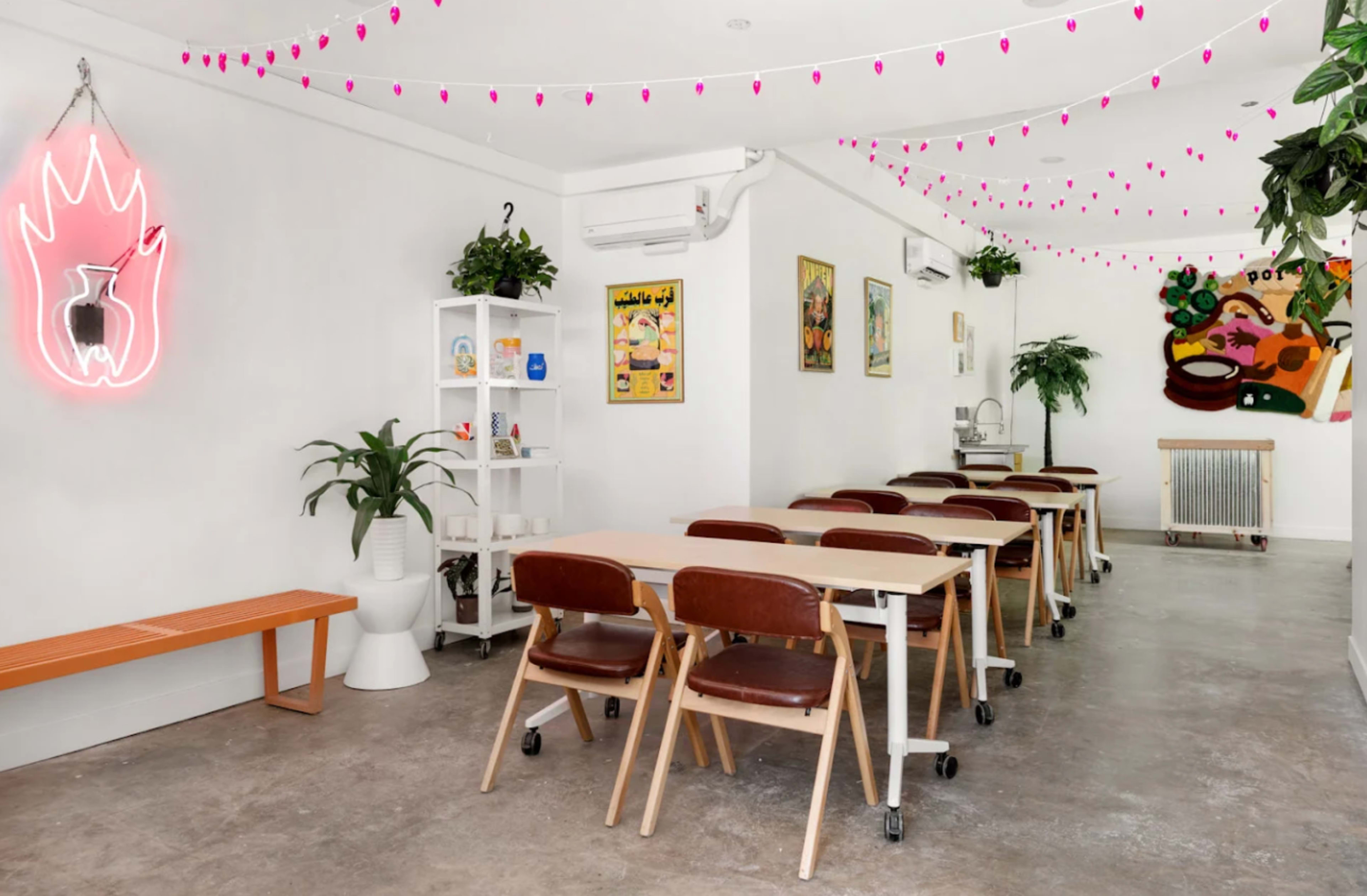 The image shows a modern, minimalist room with a long table and chairs, decorative plants, a neon light, and colorful wall art.