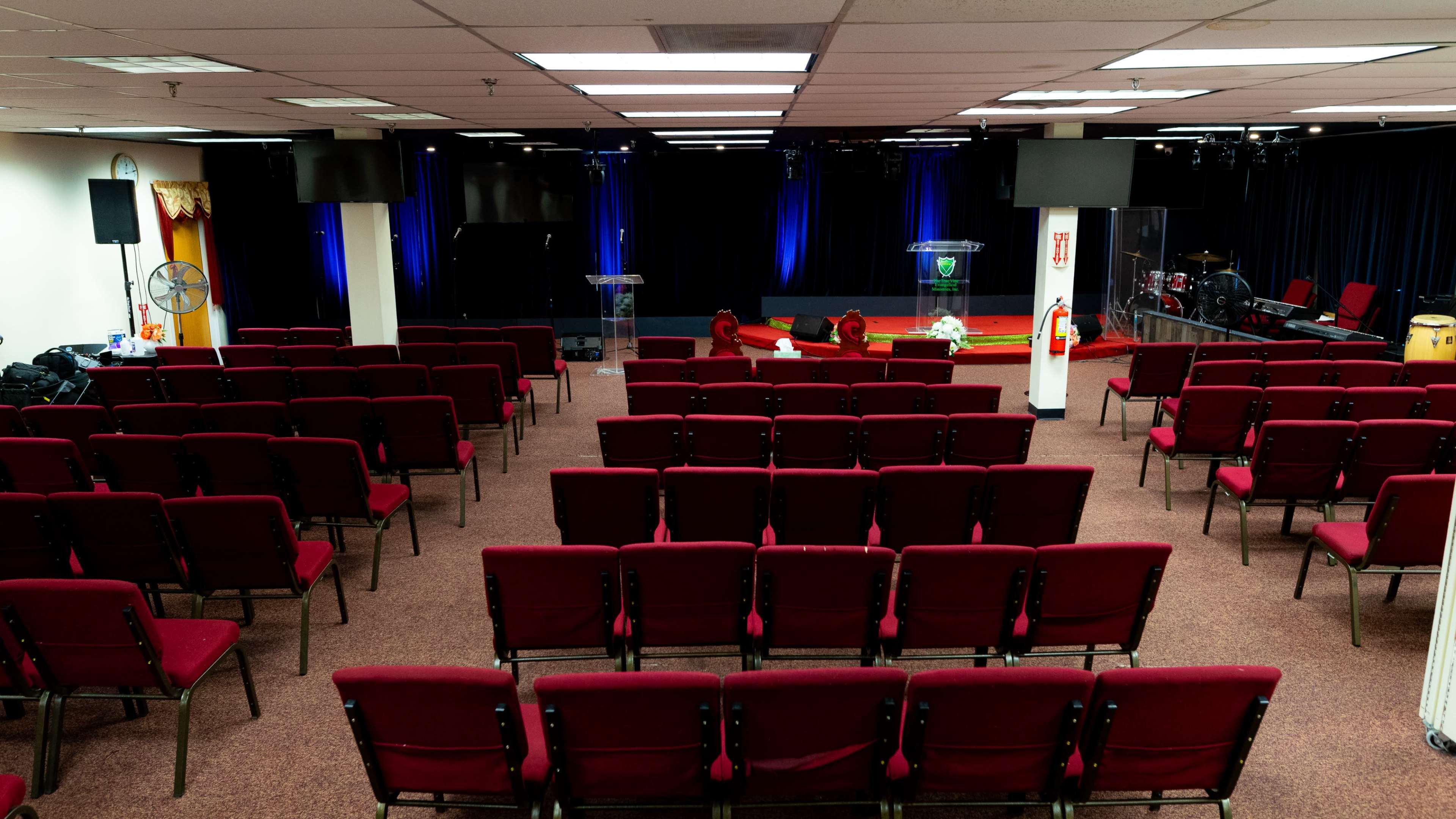 A vacant auditorium with rows of red chairs facing a stage equipped with two podiums and various musical instruments in the background.