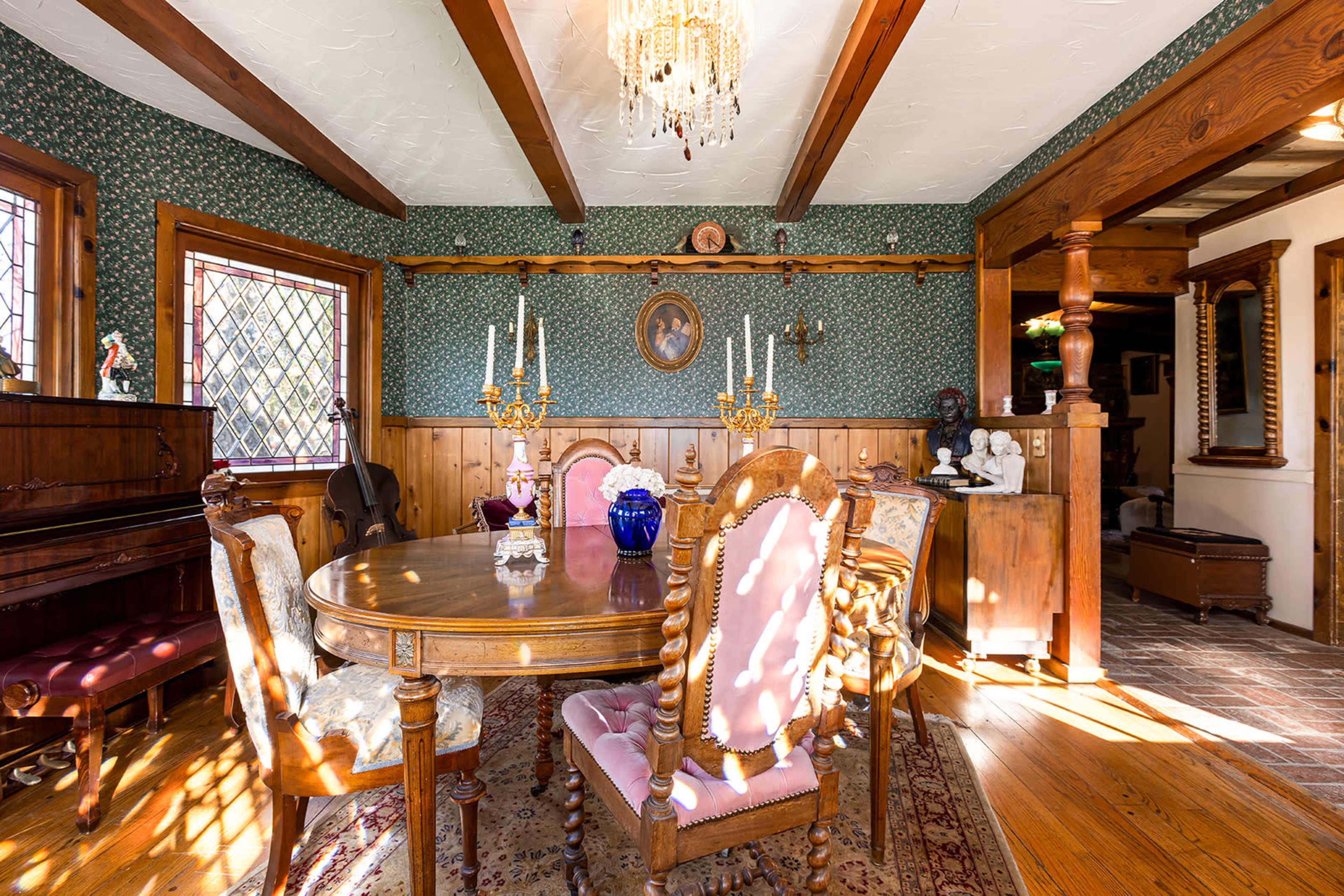 The image shows a vintage dining room featuring a wooden table surrounded by ornate chairs, a chandelier overhead, and wallpaper with floral patterns.