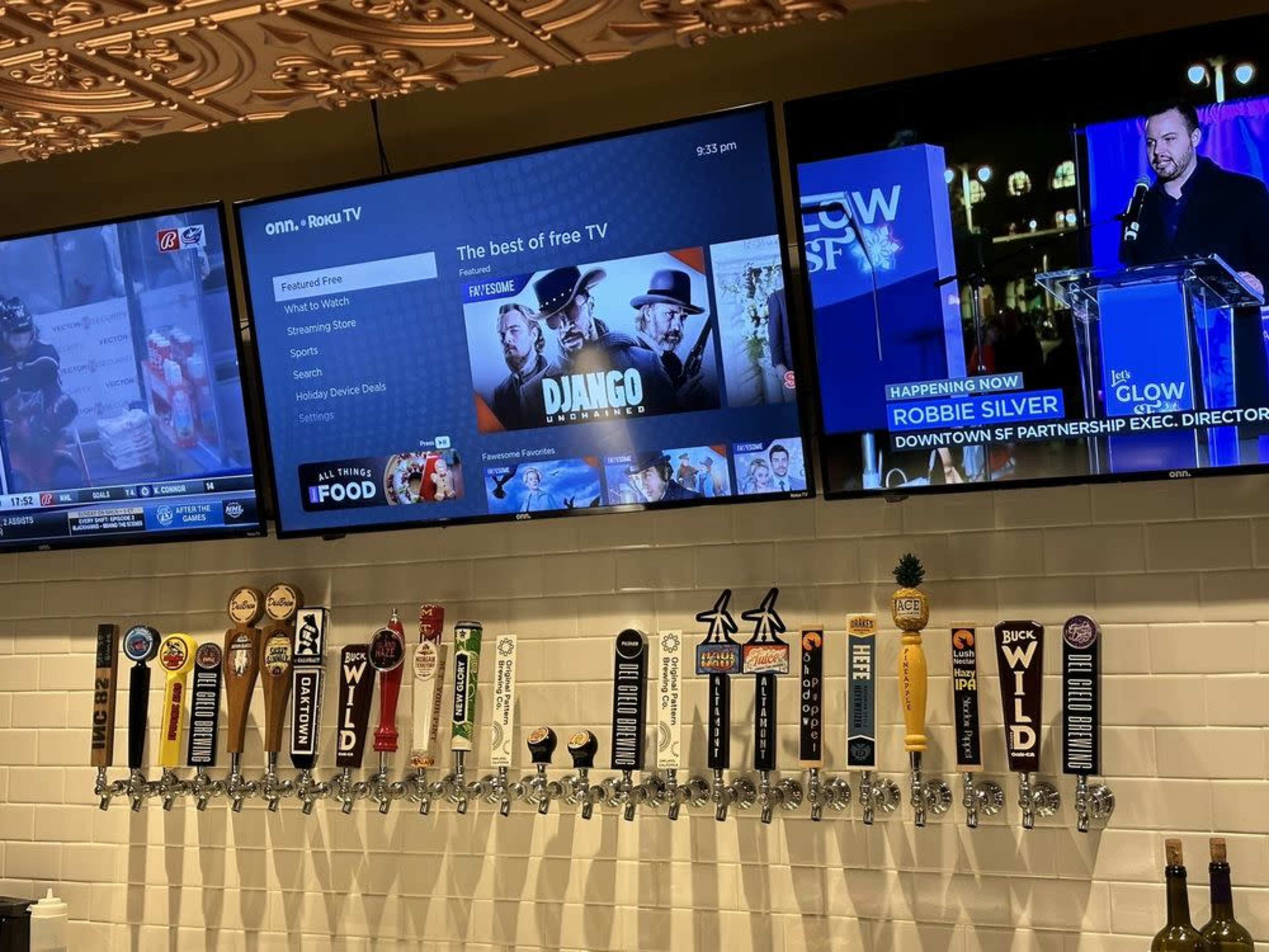 The image shows a bar area with a wall of beer tap handles and two large screens displaying different television programs.