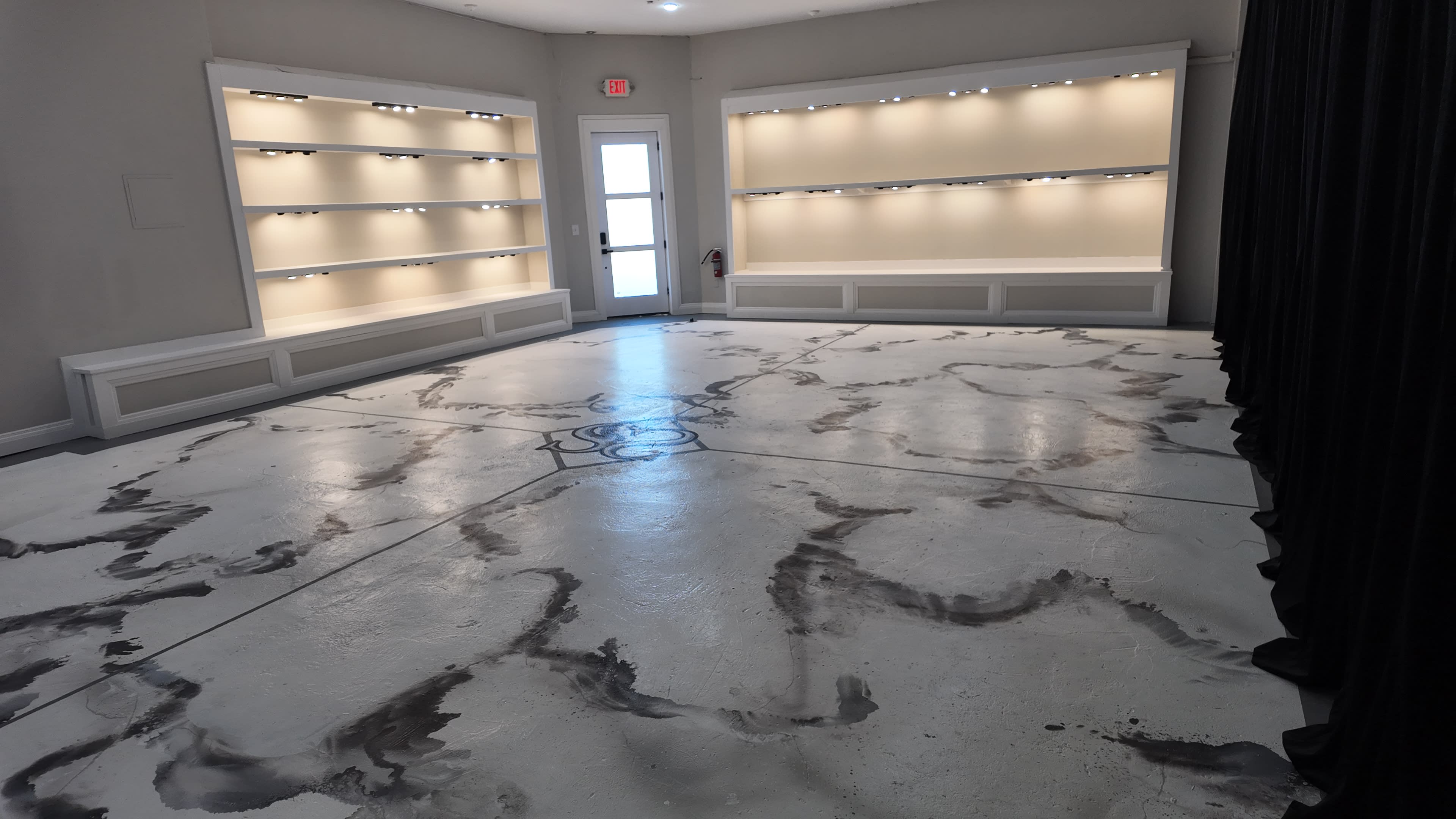 The image shows a spacious room with light-colored walls, illuminated shelves on either side, and a uniquely designed concrete floor featuring swirling gray patterns.