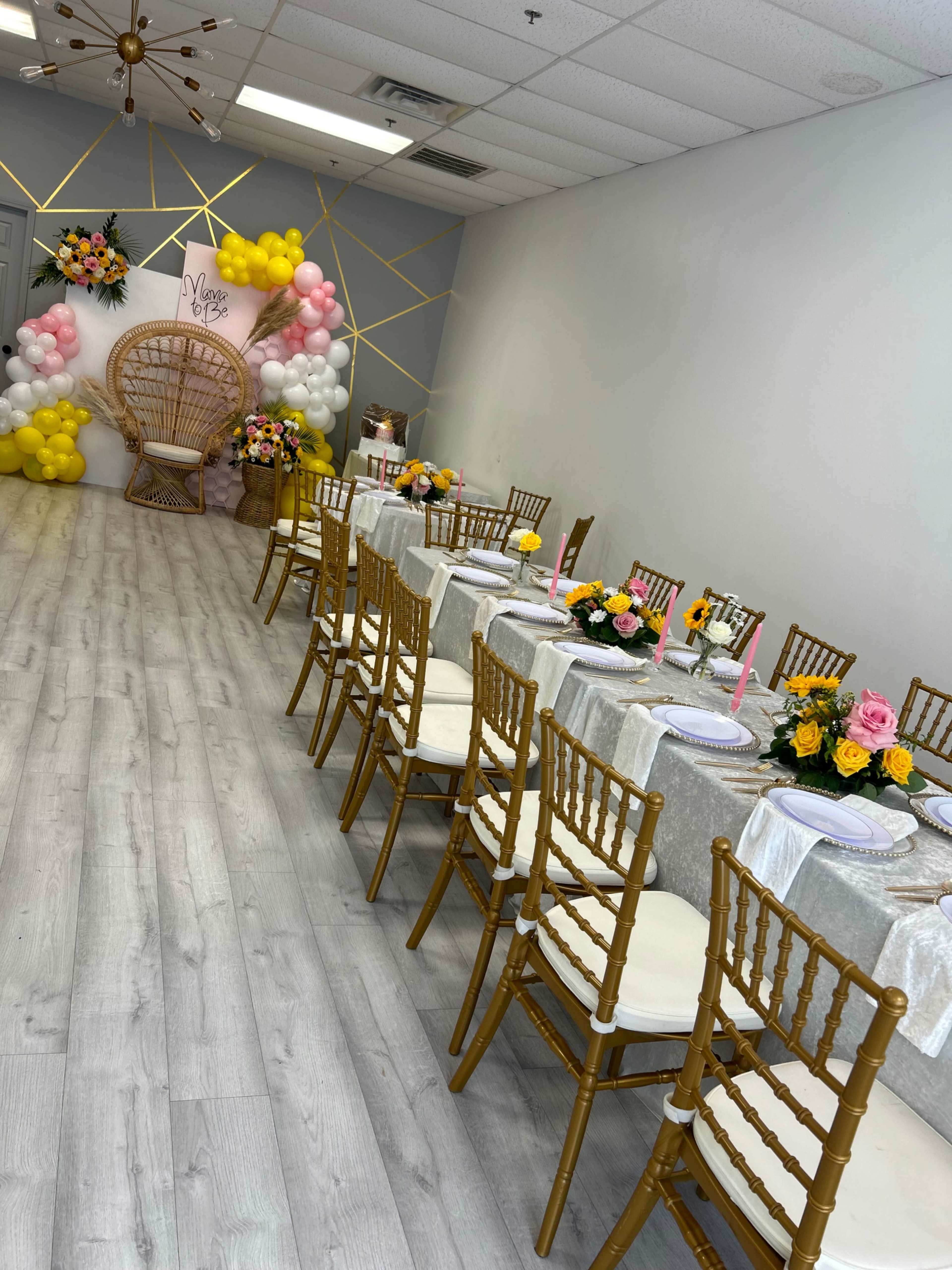 A decorated event space with rows of gold chairs, tables set with floral centerpieces, and a backdrop featuring balloons and a large wicker chair.