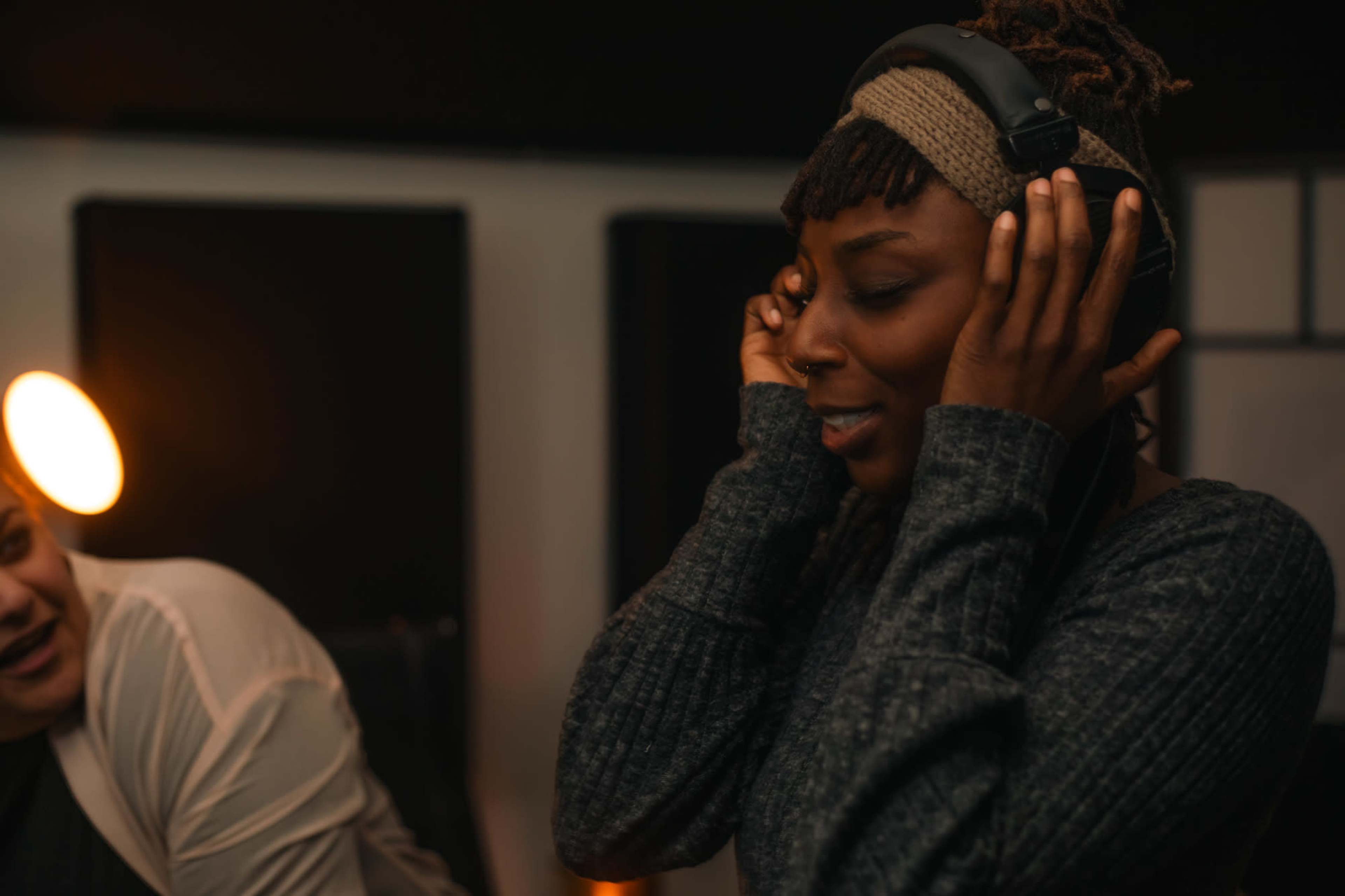 A woman wearing headphones listens intently while another person is partially visible in the background.