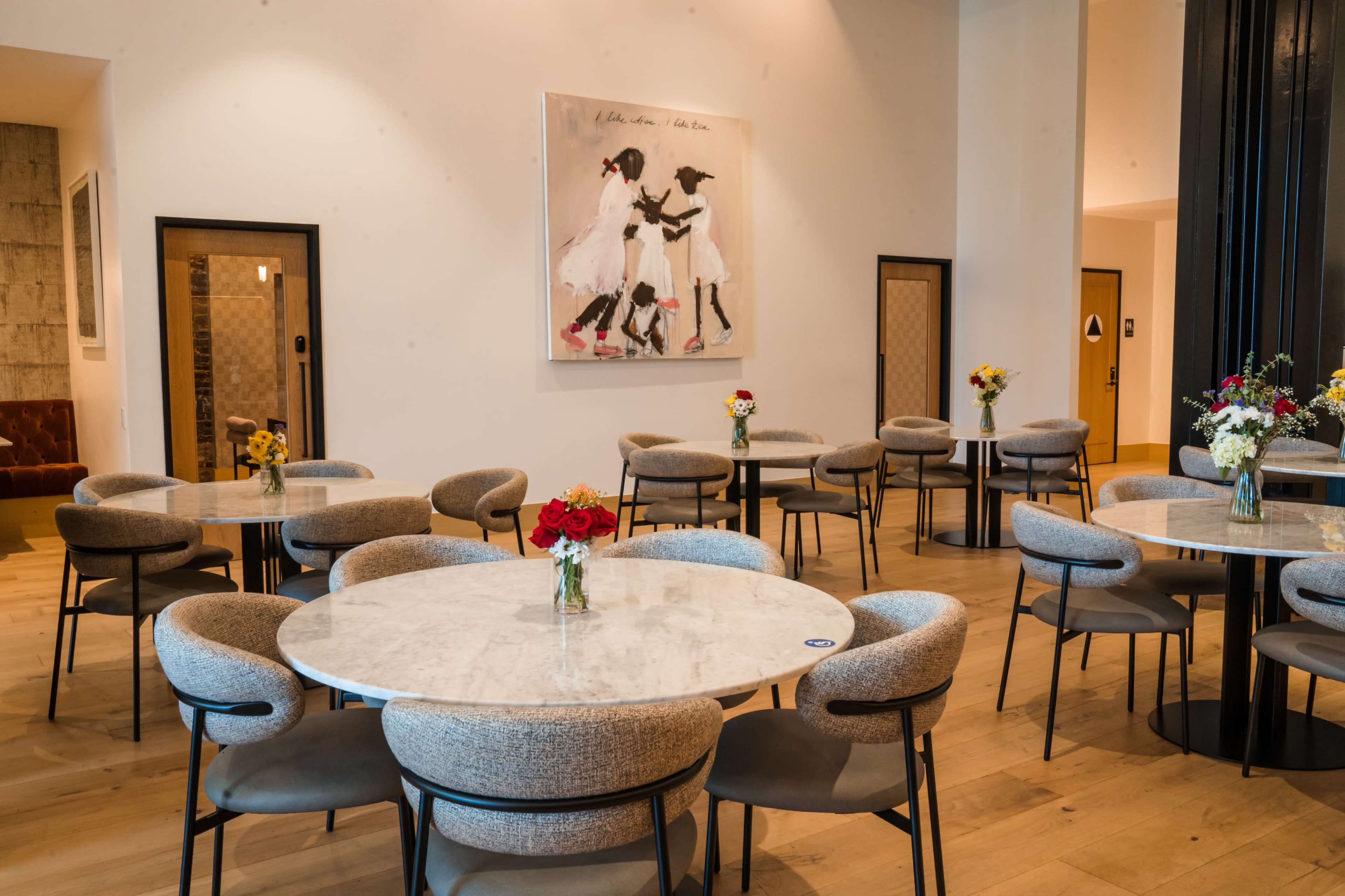 A modern dining area with round marble tables, upholstered chairs, and small flower arrangements on each table, against a backdrop of a large framed artwork.