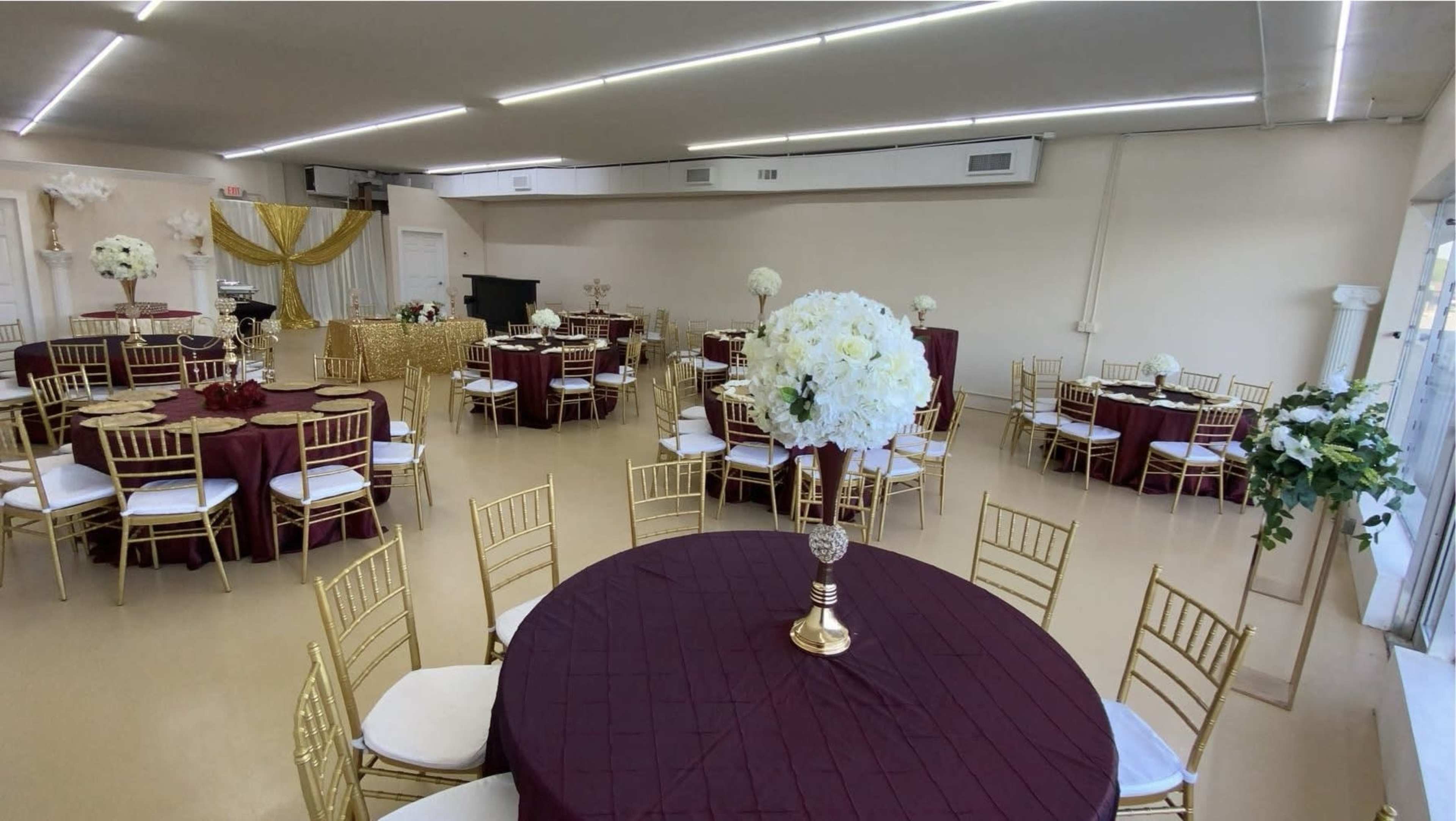 A banquet hall set up with round tables covered in maroon tablecloths and adorned with gold chairs and floral centerpieces.