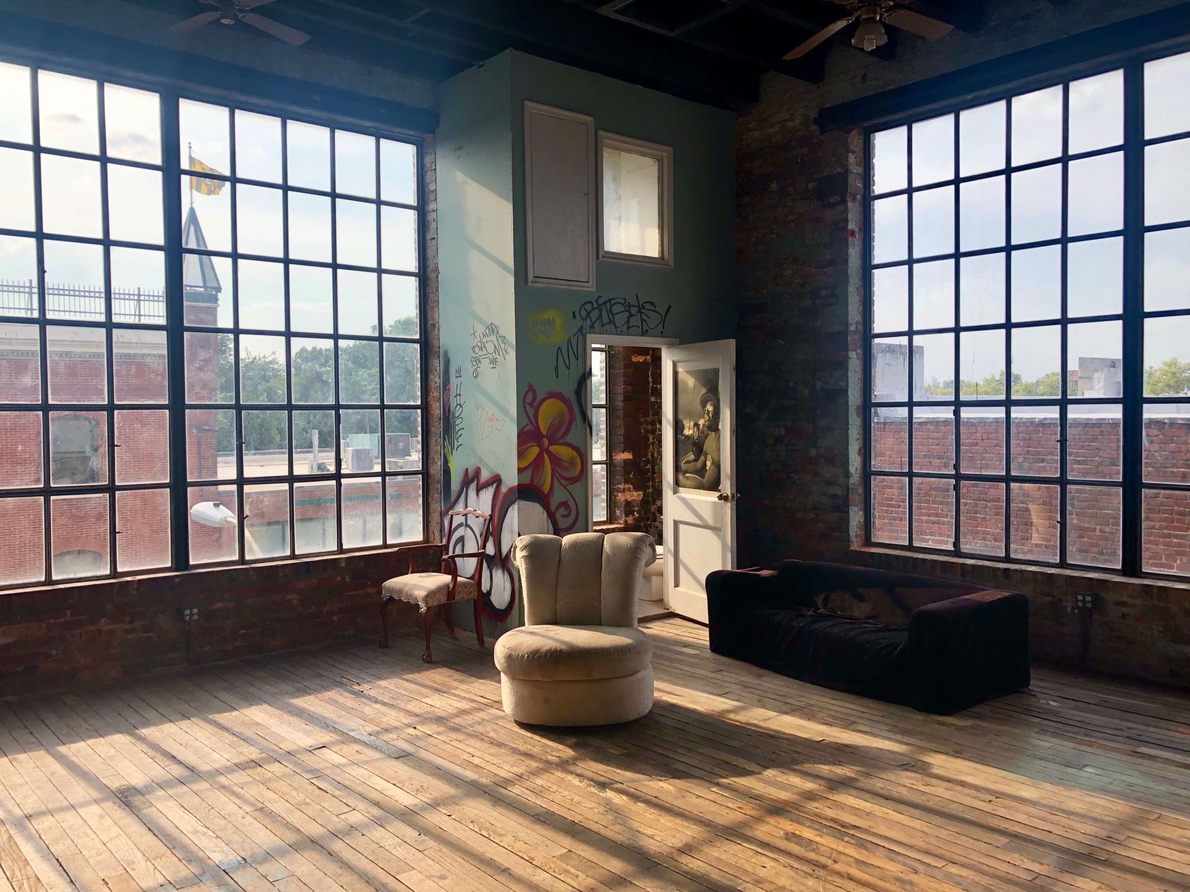 A spacious, industrial-style room features large windows, exposed brick walls, and minimal furnishings, including a round chair and a dark couch.