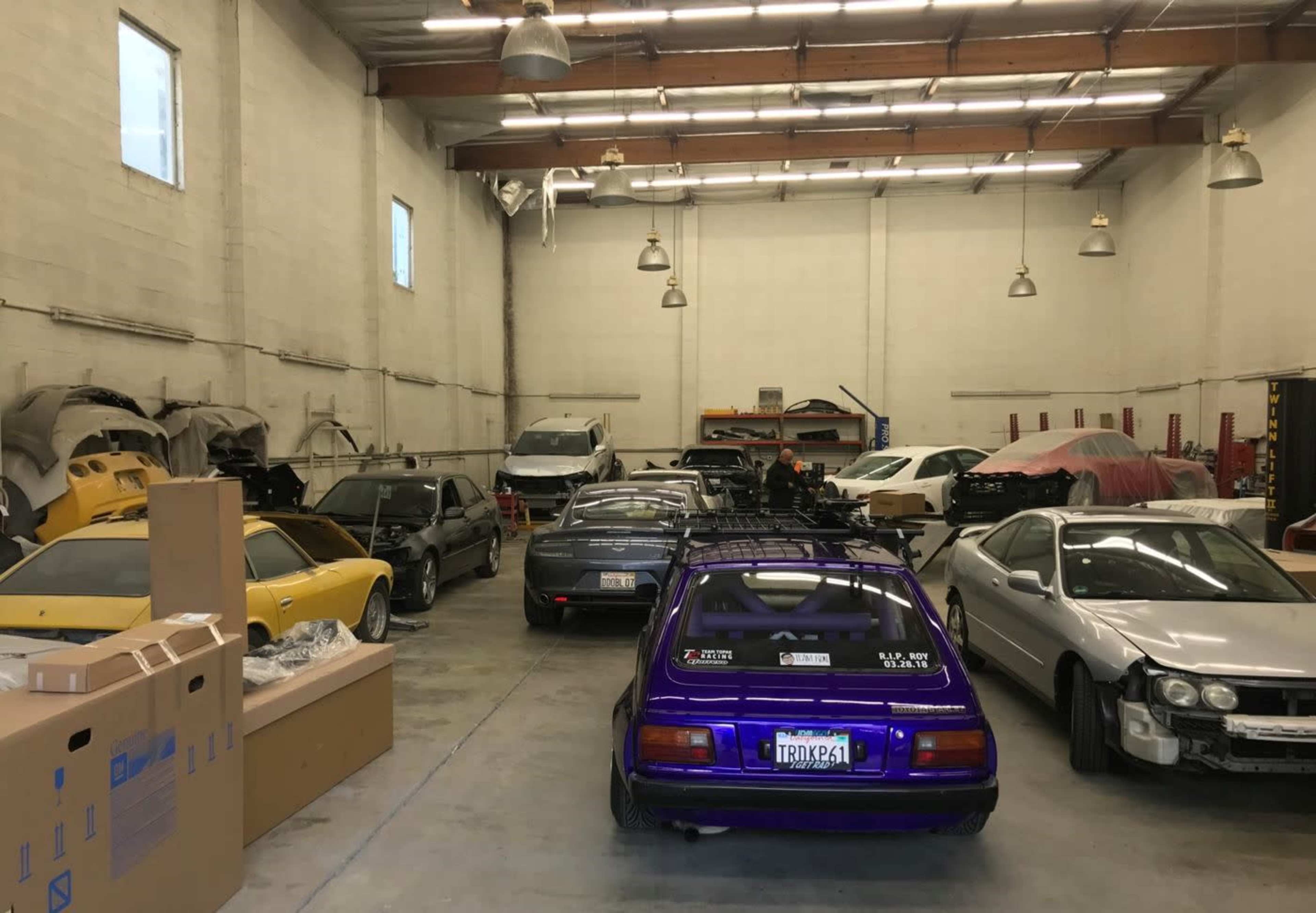 The image shows a garage filled with various cars, including a purple vehicle in the foreground, and several others covered or partially visible in a spacious, well-lit interior.