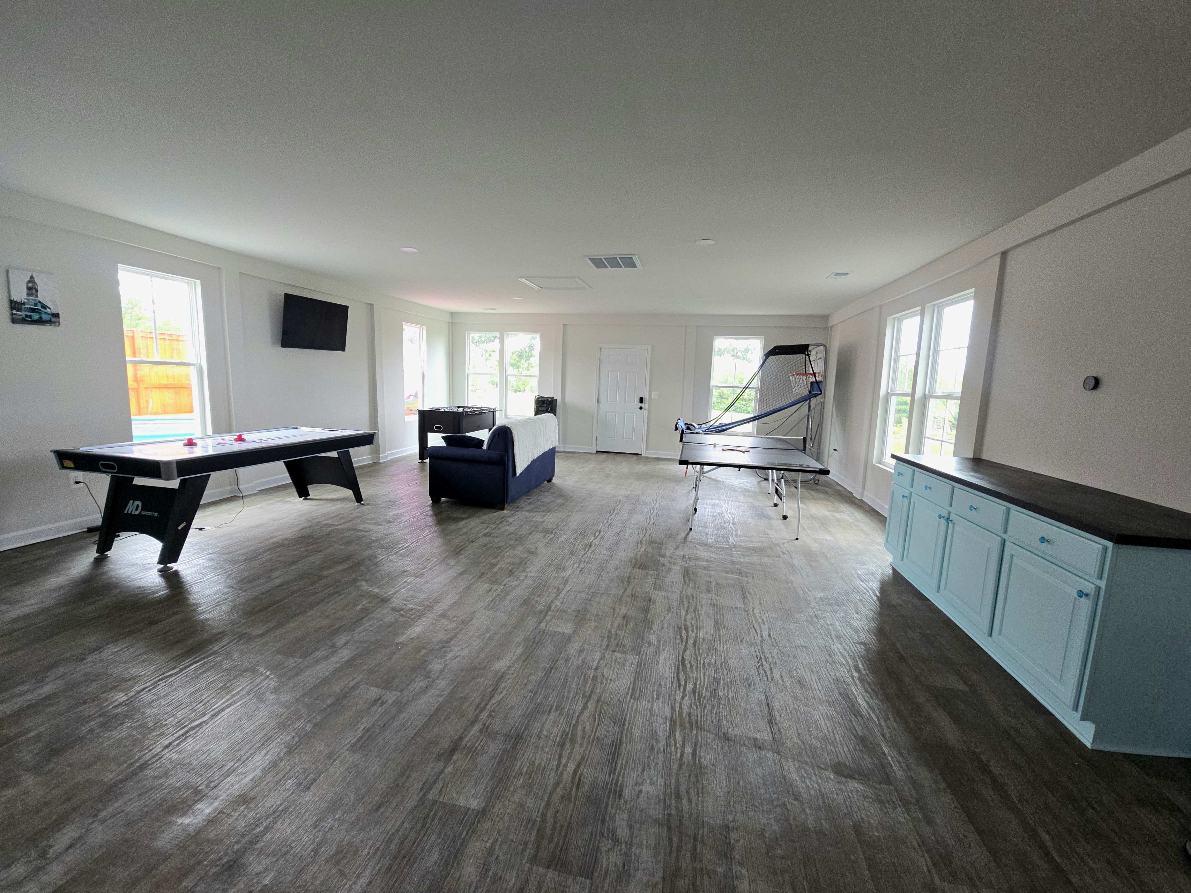 The image shows a spacious, well-lit room featuring a pool table, a table tennis setup, a couch, and large windows.