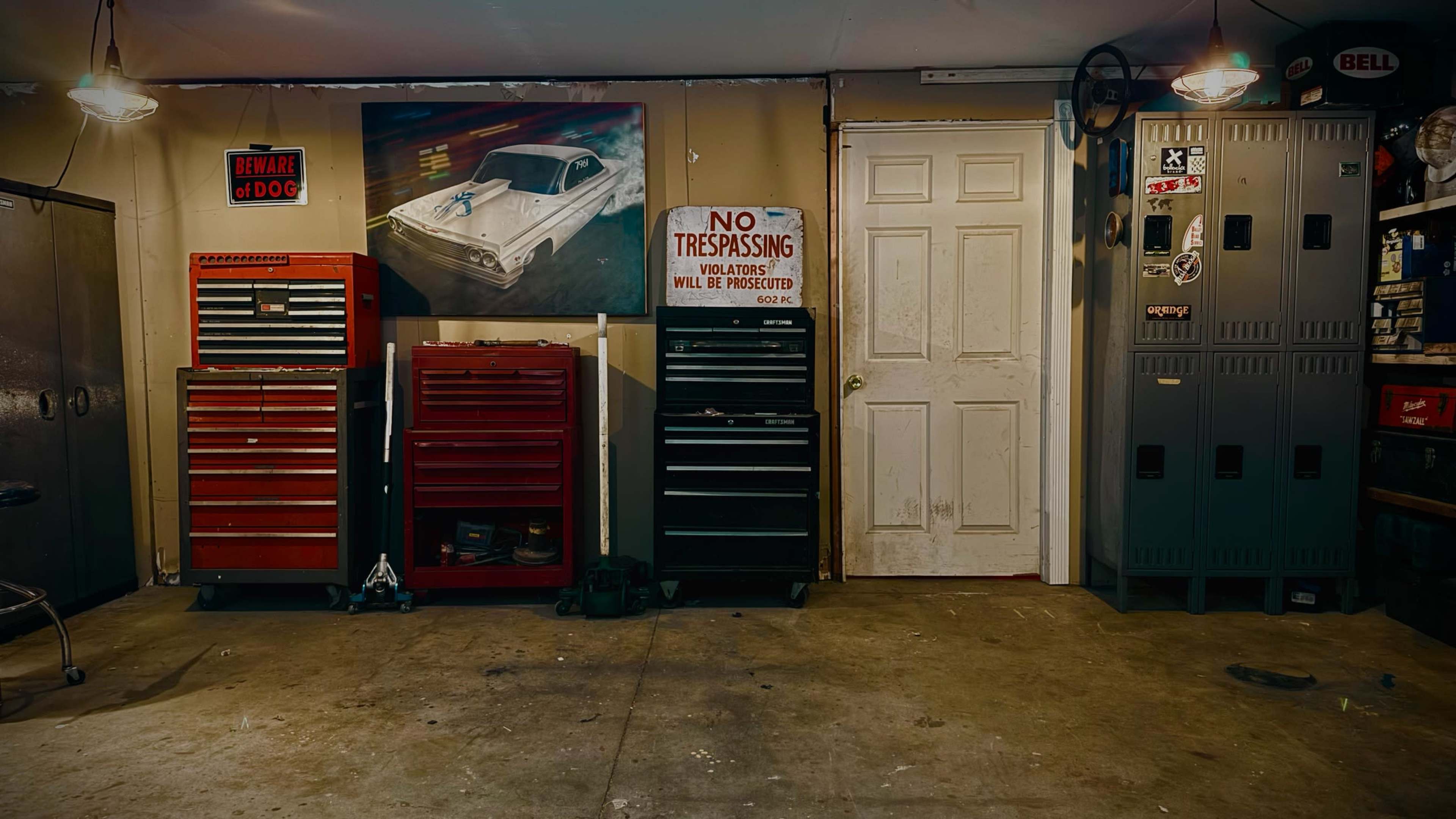 The image shows a garage interior with two tall tool chests in red and black, a metal cabinet, and a door leading outside, all arranged against a wall featuring a car painting and a "No Trespassing" sign.