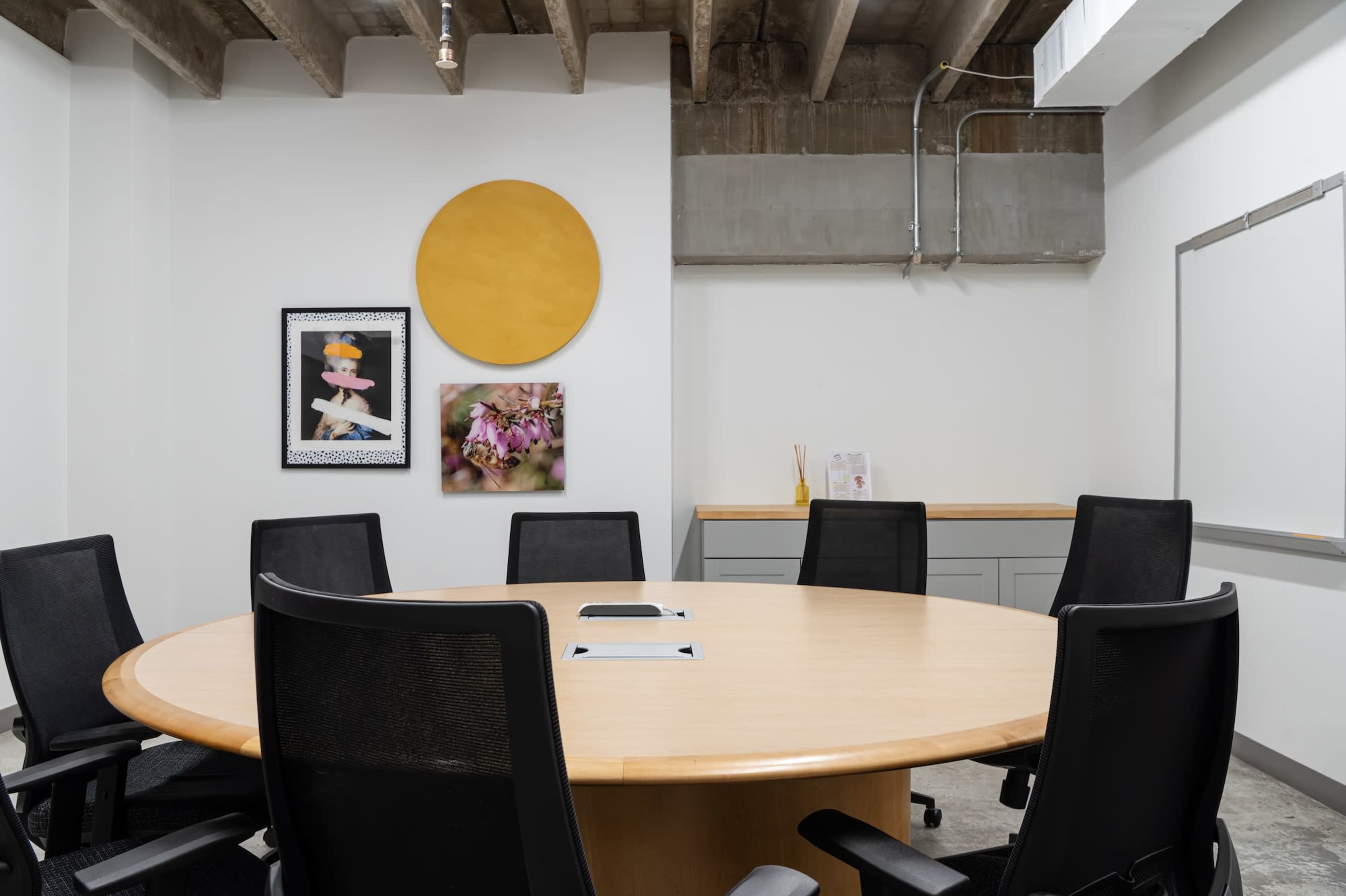 The image shows a modern conference room with a large round table, surrounded by black chairs, and features two framed artworks on the wall alongside a circular wall decoration.