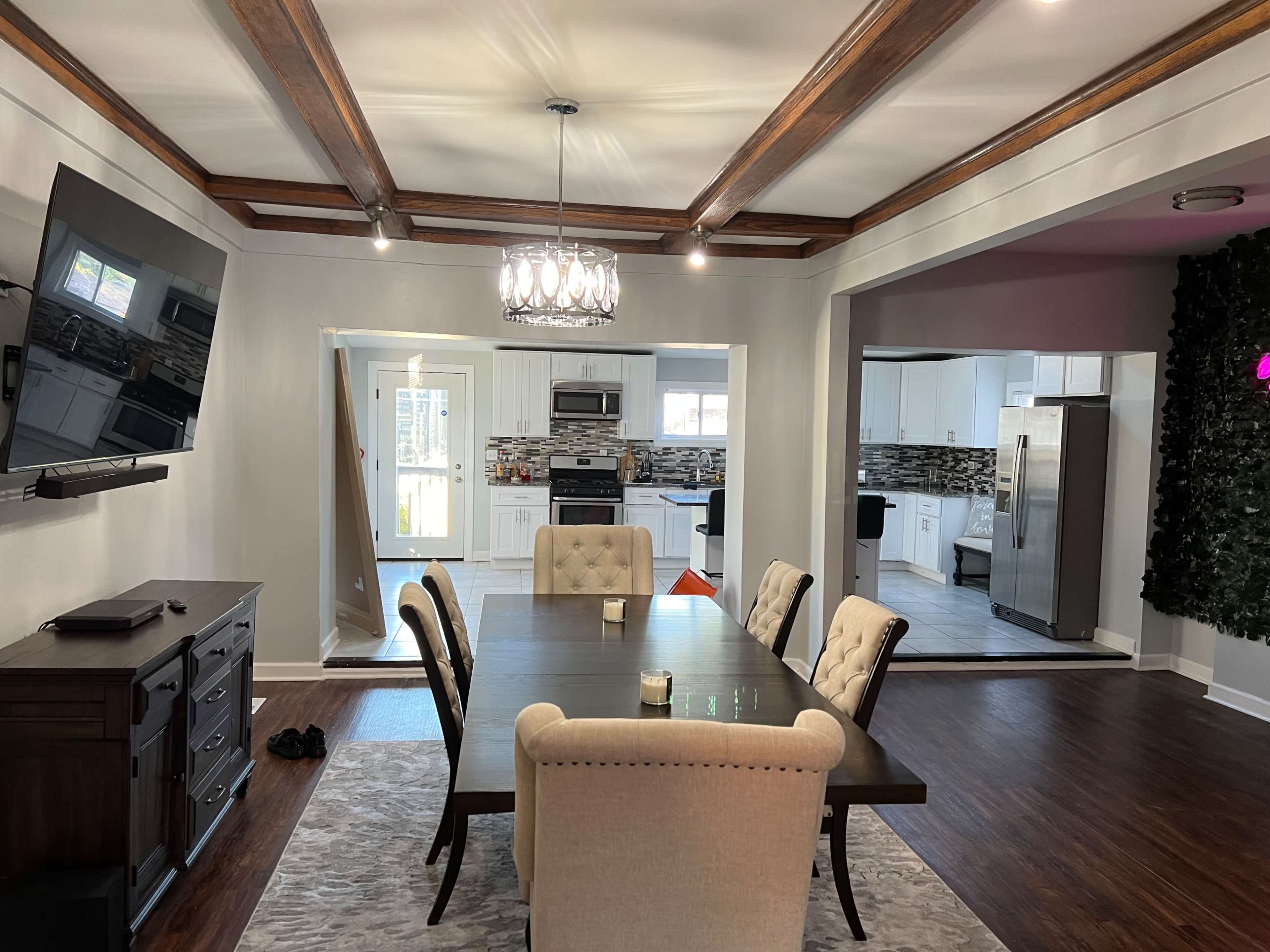 A dining area with a dark wooden table surrounded by upholstered chairs, a chandelier overhead, and an open view into a kitchen with modern appliances and decor.