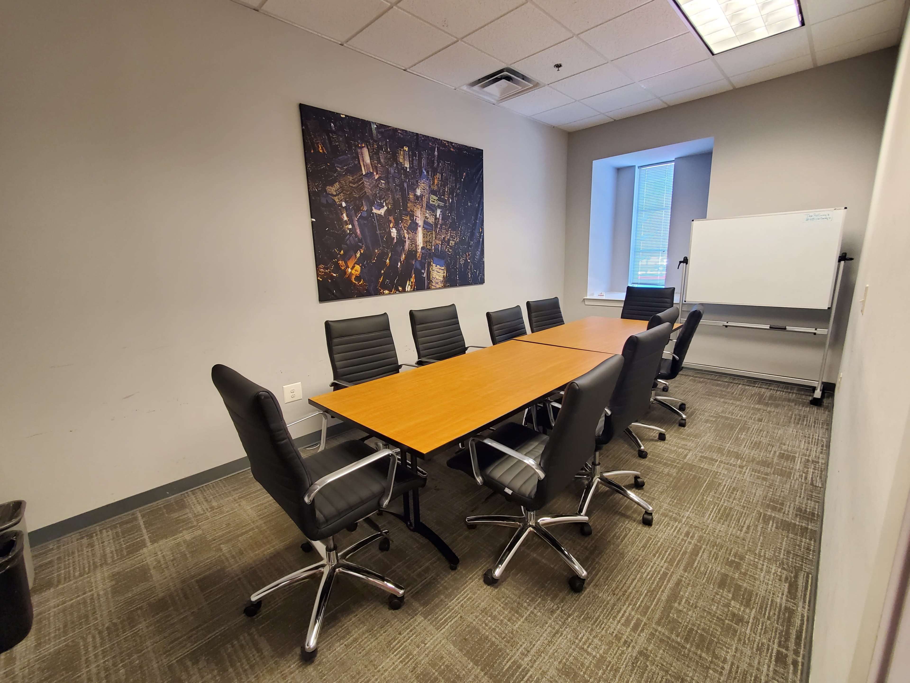 A meeting room features a rectangular arrangement of wooden tables surrounded by black leather chairs, with a large cityscape artwork on the wall and a whiteboard in one corner.
