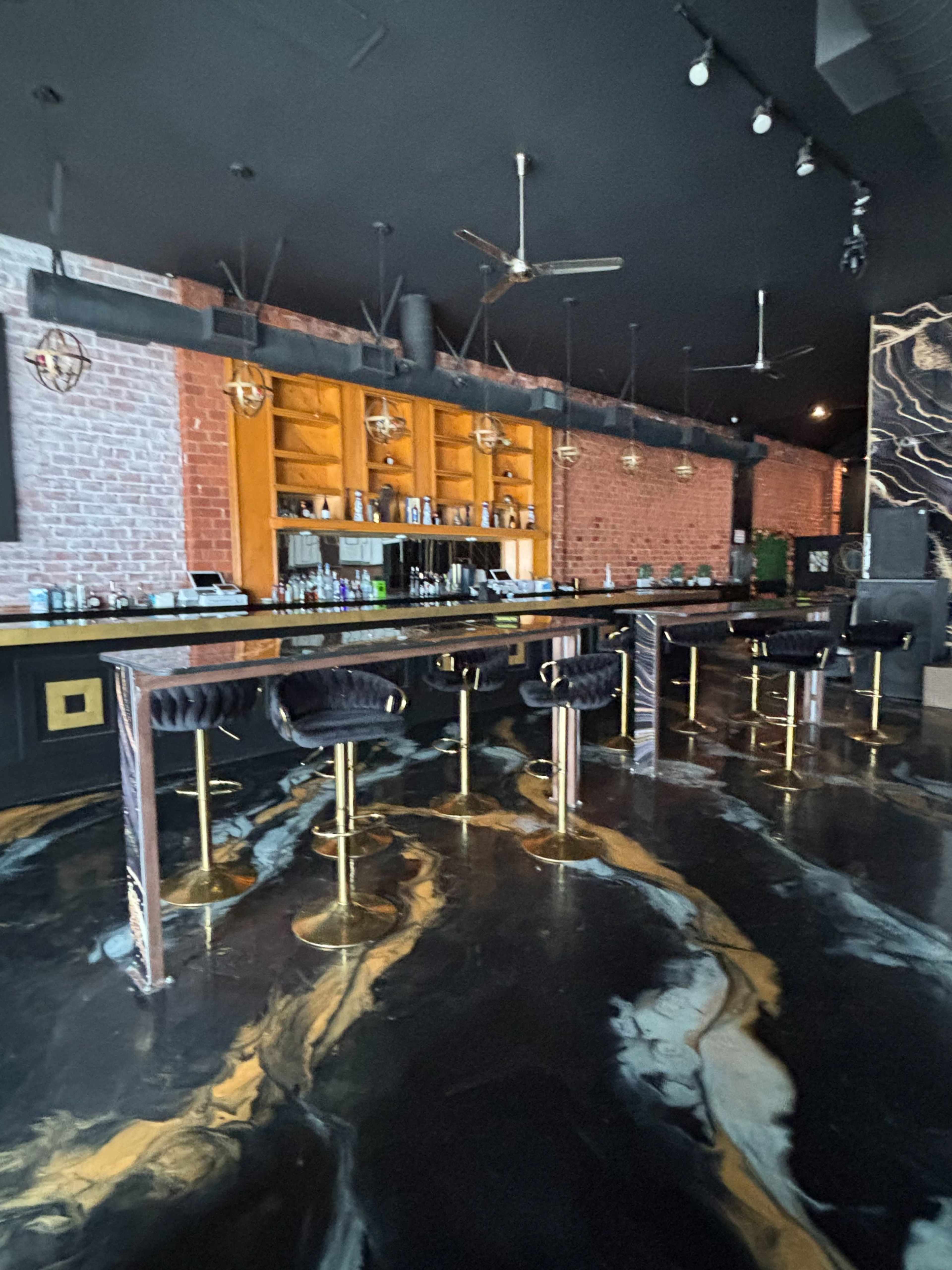 The image shows a modern bar interior with a black and gold floor, exposed brick walls, and a sleek bar with high stools.