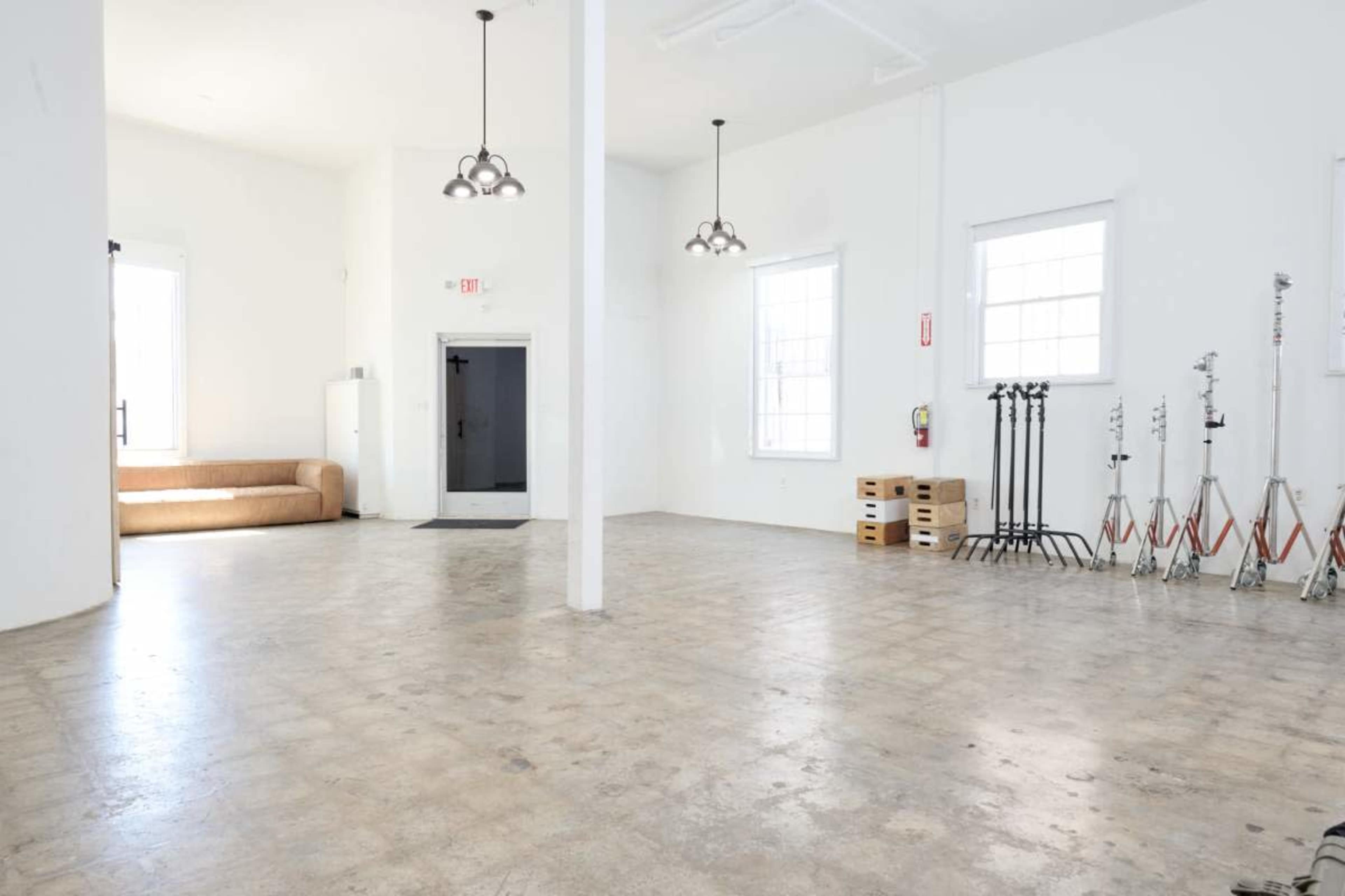 The image shows an empty room with white walls, large windows, a couch to the left, and equipment stands on the right.