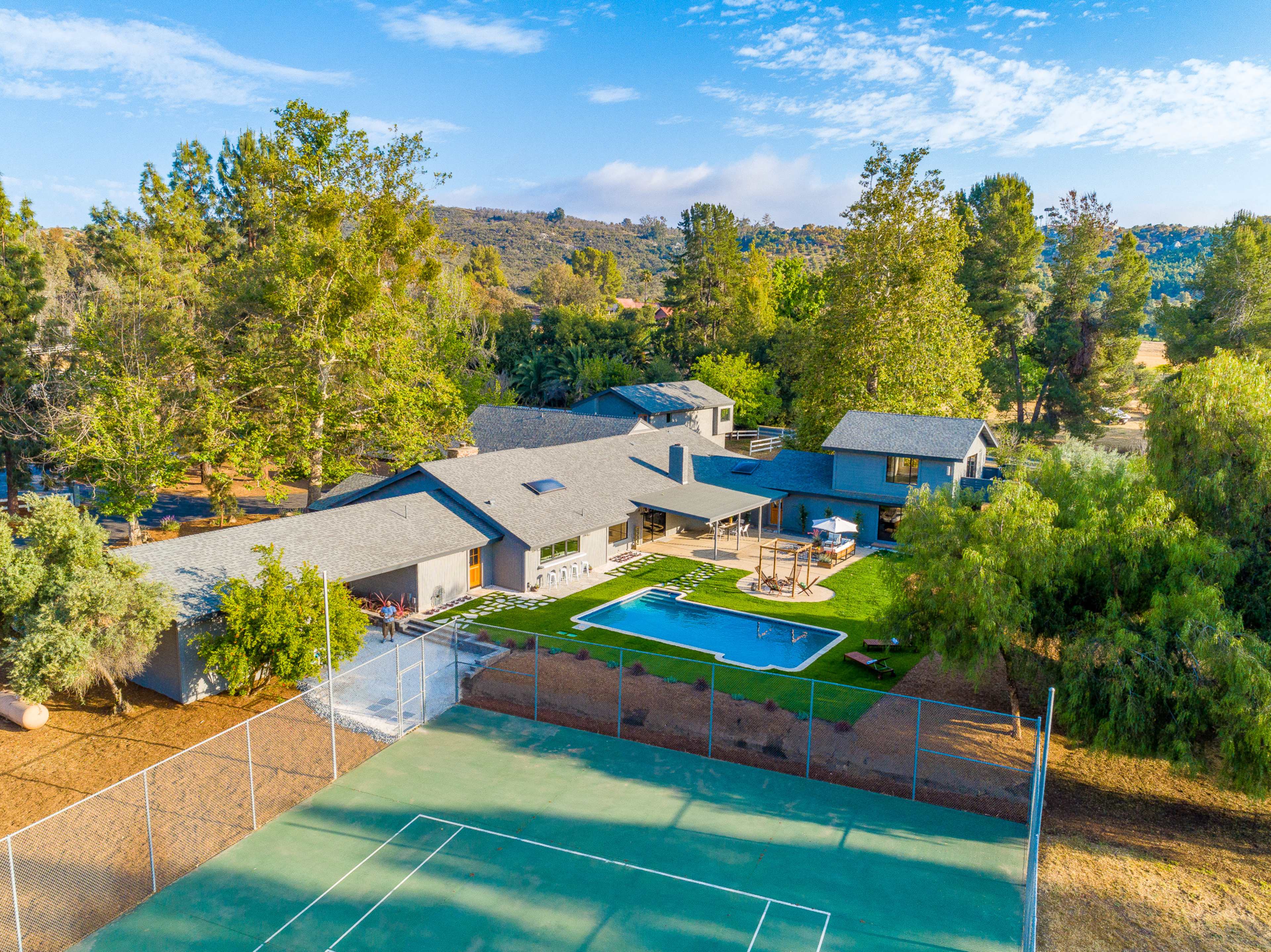 The image shows a modern house with a swimming pool and tennis court, surrounded by lush greenery and trees.