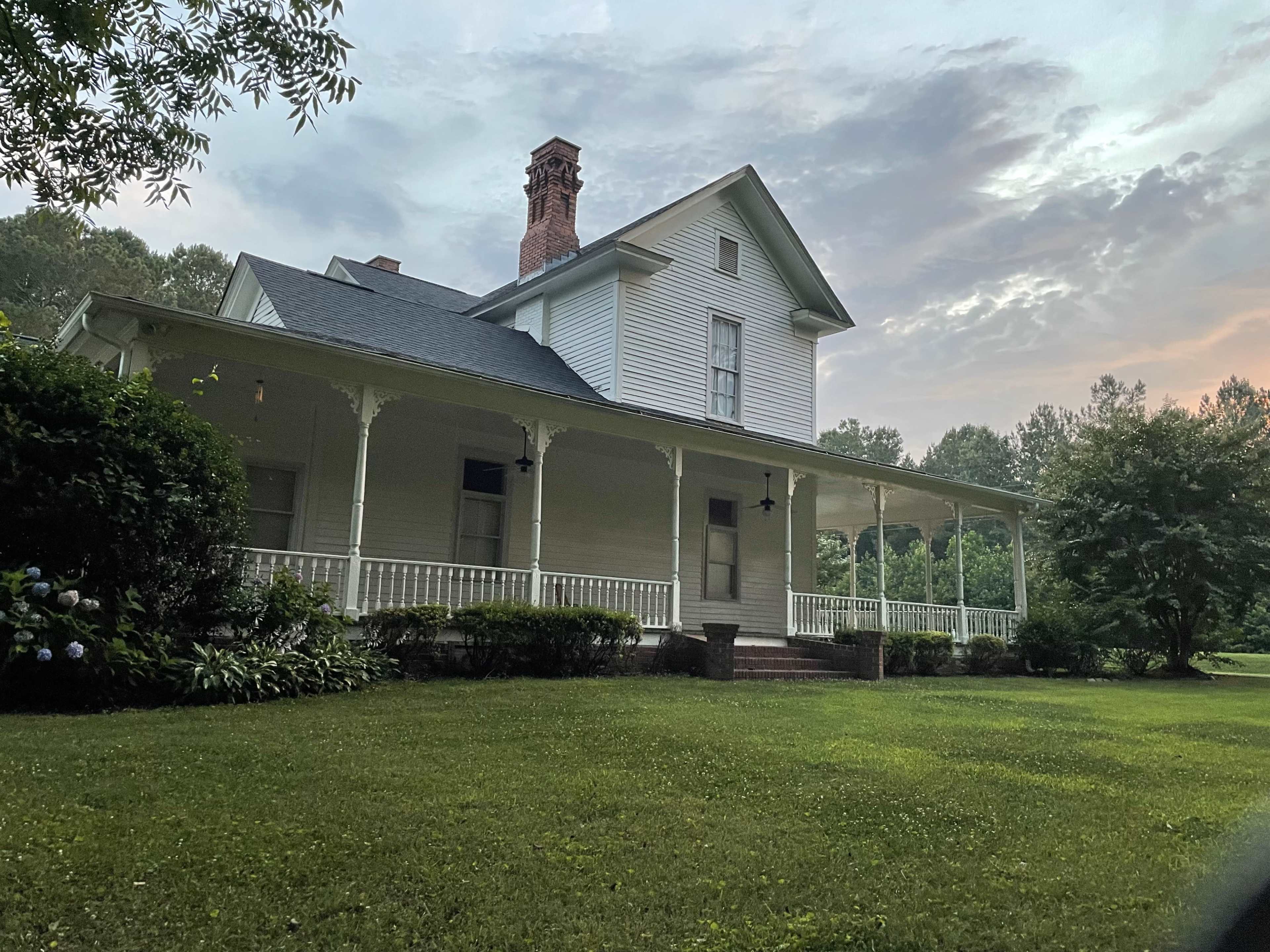 A white two-story house with a front porch and a chimney is set in a grassy area surrounded by trees.