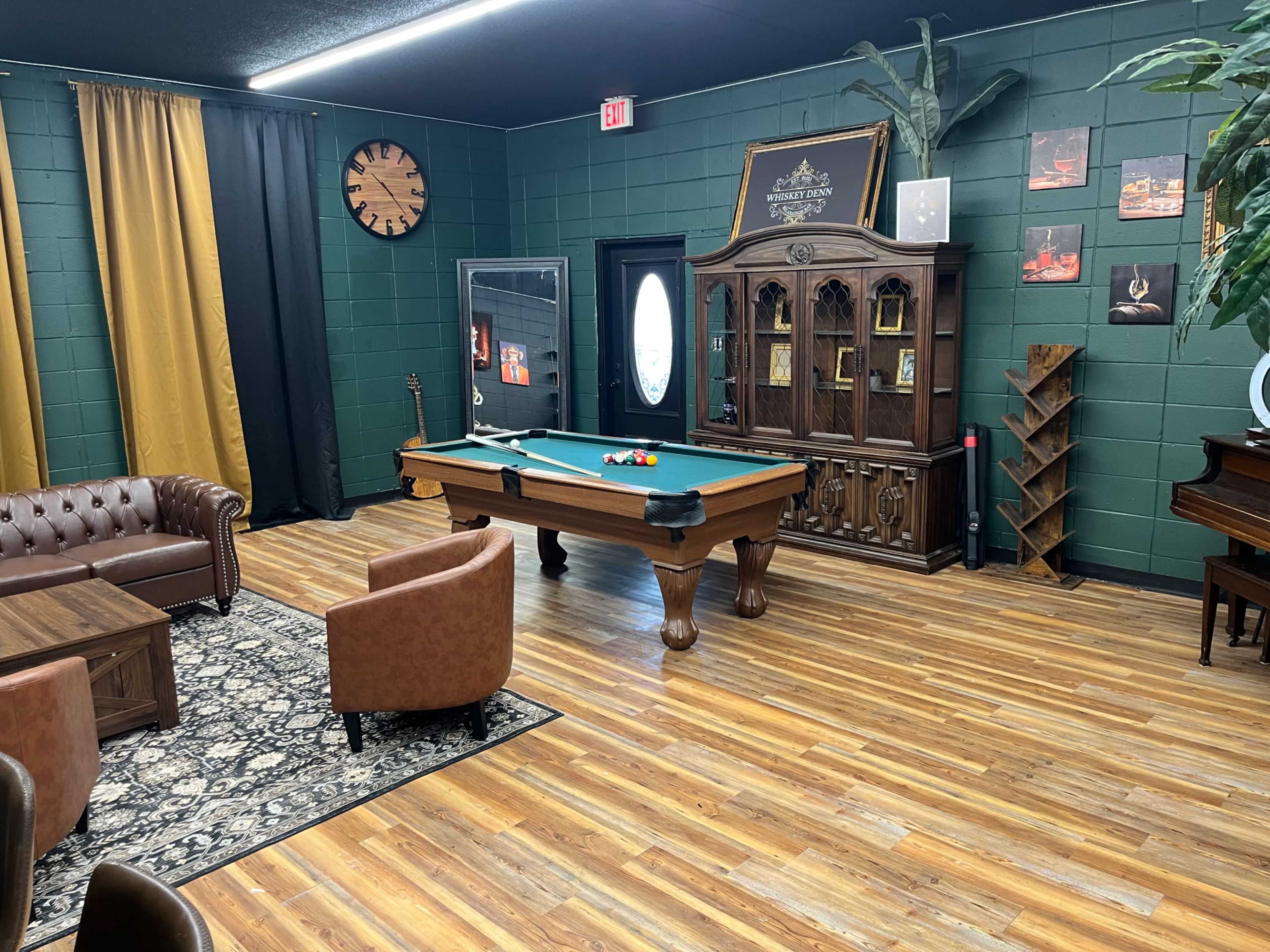 The image shows a recreational room with a pool table, two armchairs, a wooden table, and a decorative cabinet against a green wall, complemented by wooden flooring.