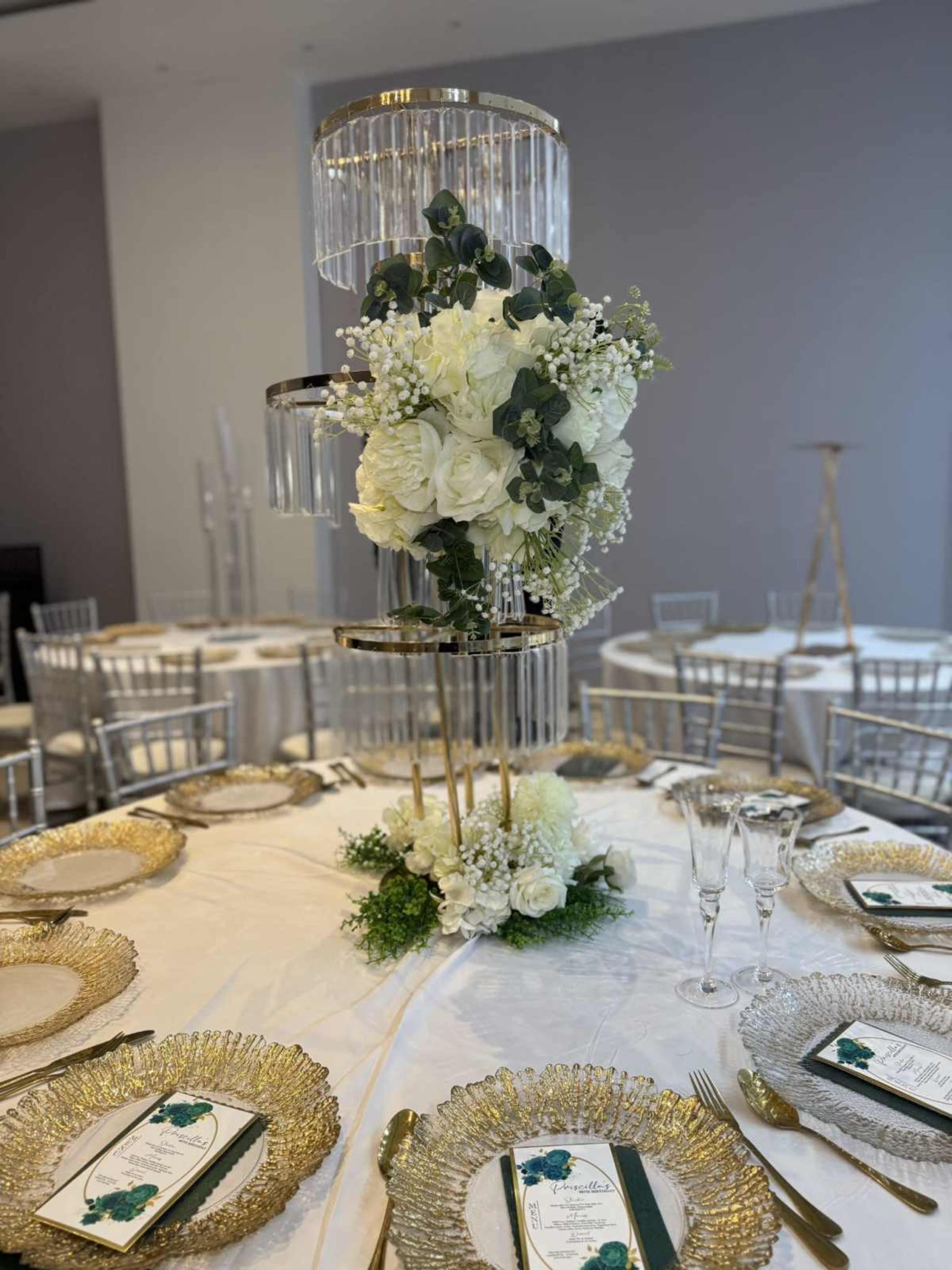 The image shows an elegantly set dining table with gold-rimmed plates, crystal glasses, and a tall floral centerpiece featuring white flowers and greenery.