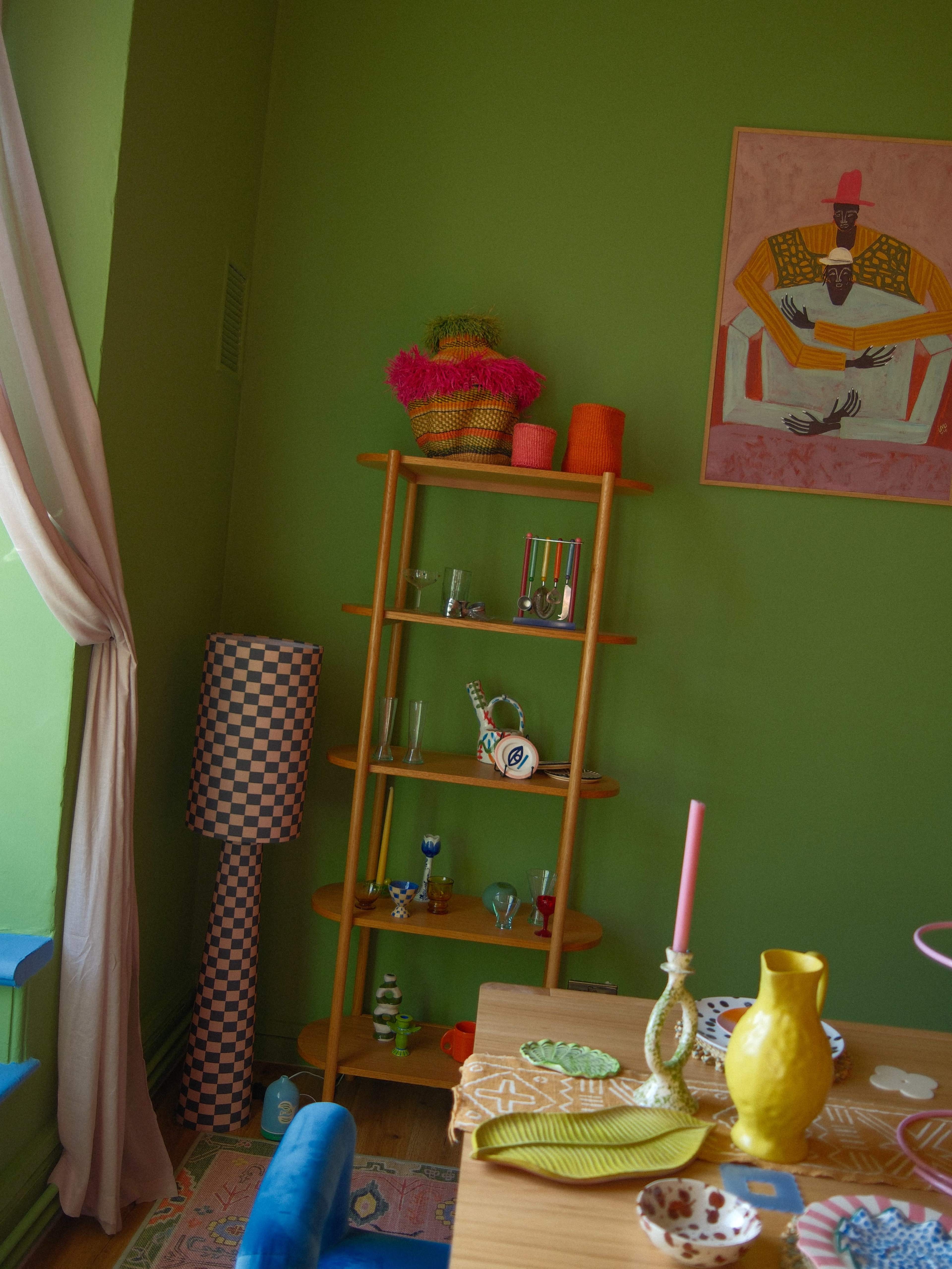 The image shows a vibrant green room featuring a wooden shelf with colorful decorative items, a checkered lamp, and a dining table set with various dishes and a pink candle.