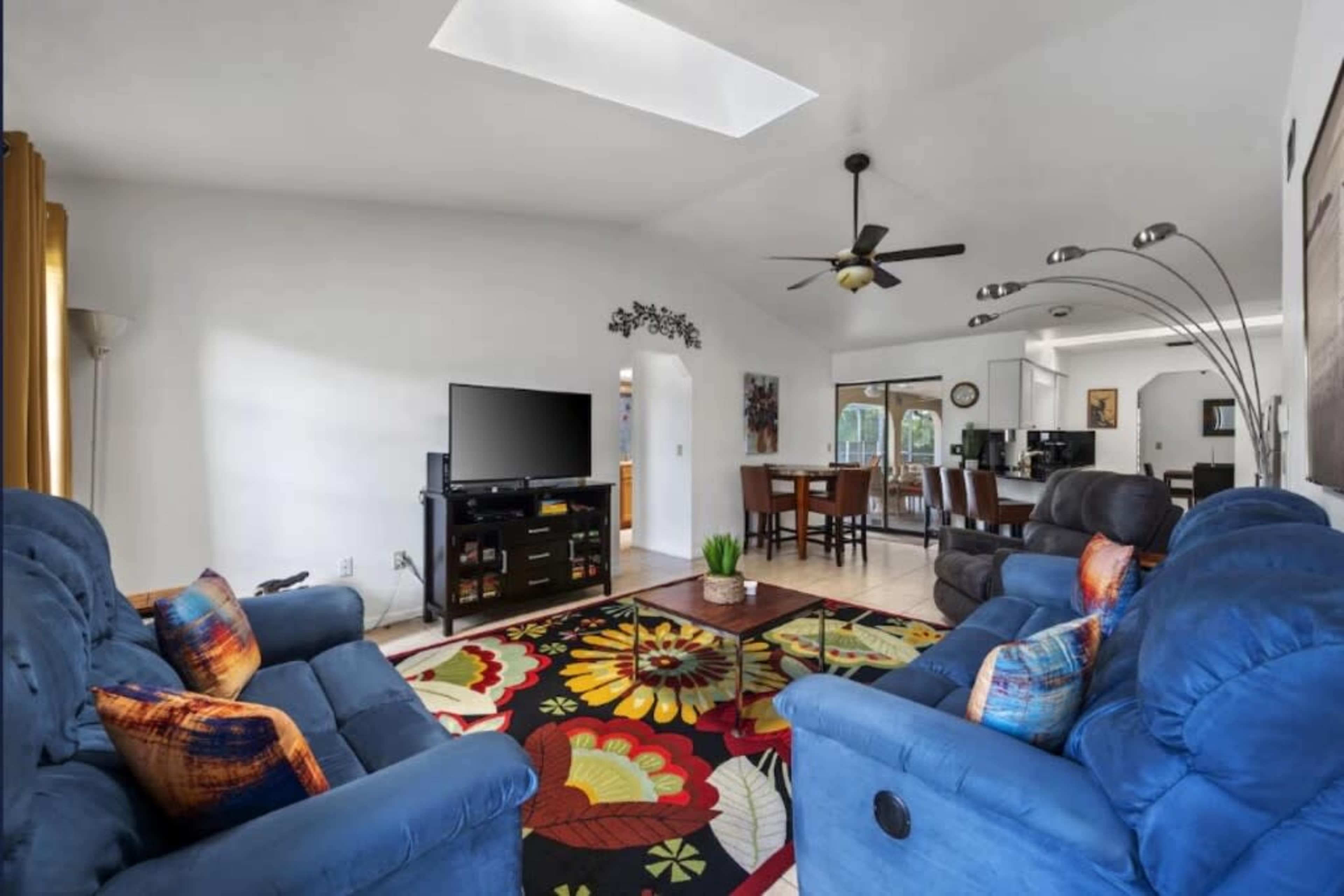 The image shows a living room with two blue sofas, a colorful rug, a television setup, and a dining area in the background.