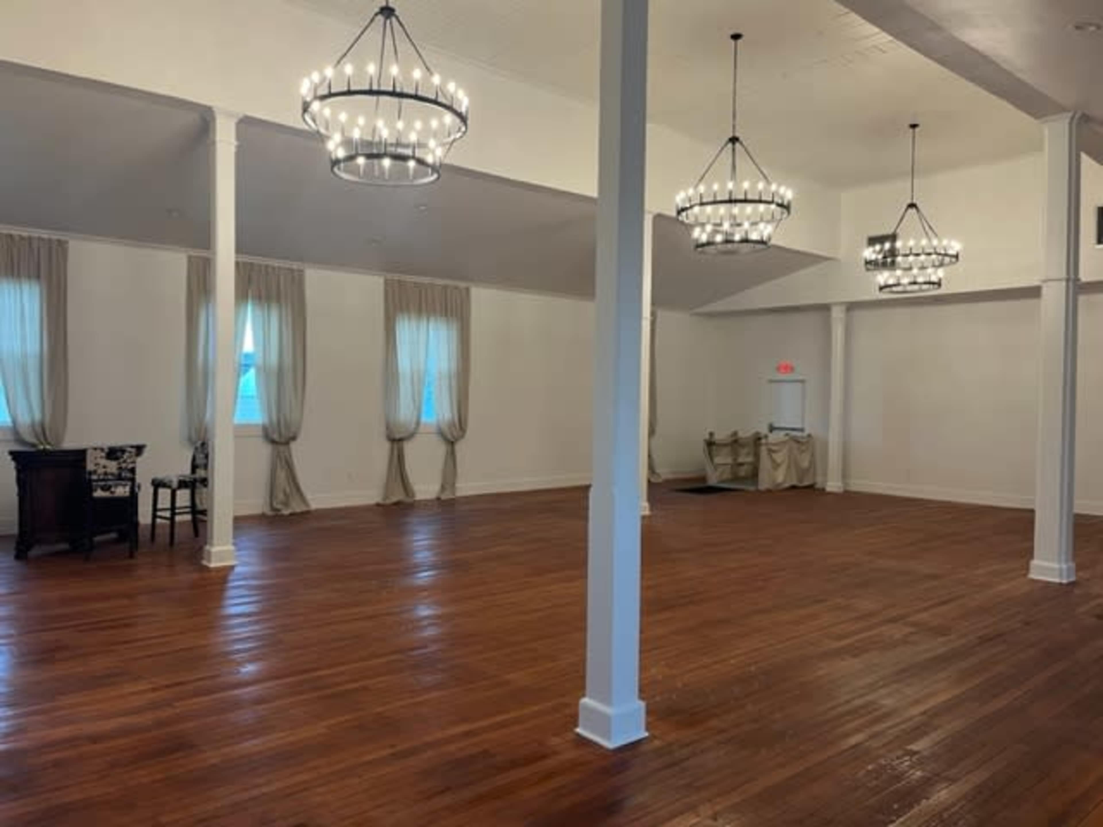 A spacious, well-lit room with wooden flooring, two elegant chandeliers, and draped curtains at the windows.