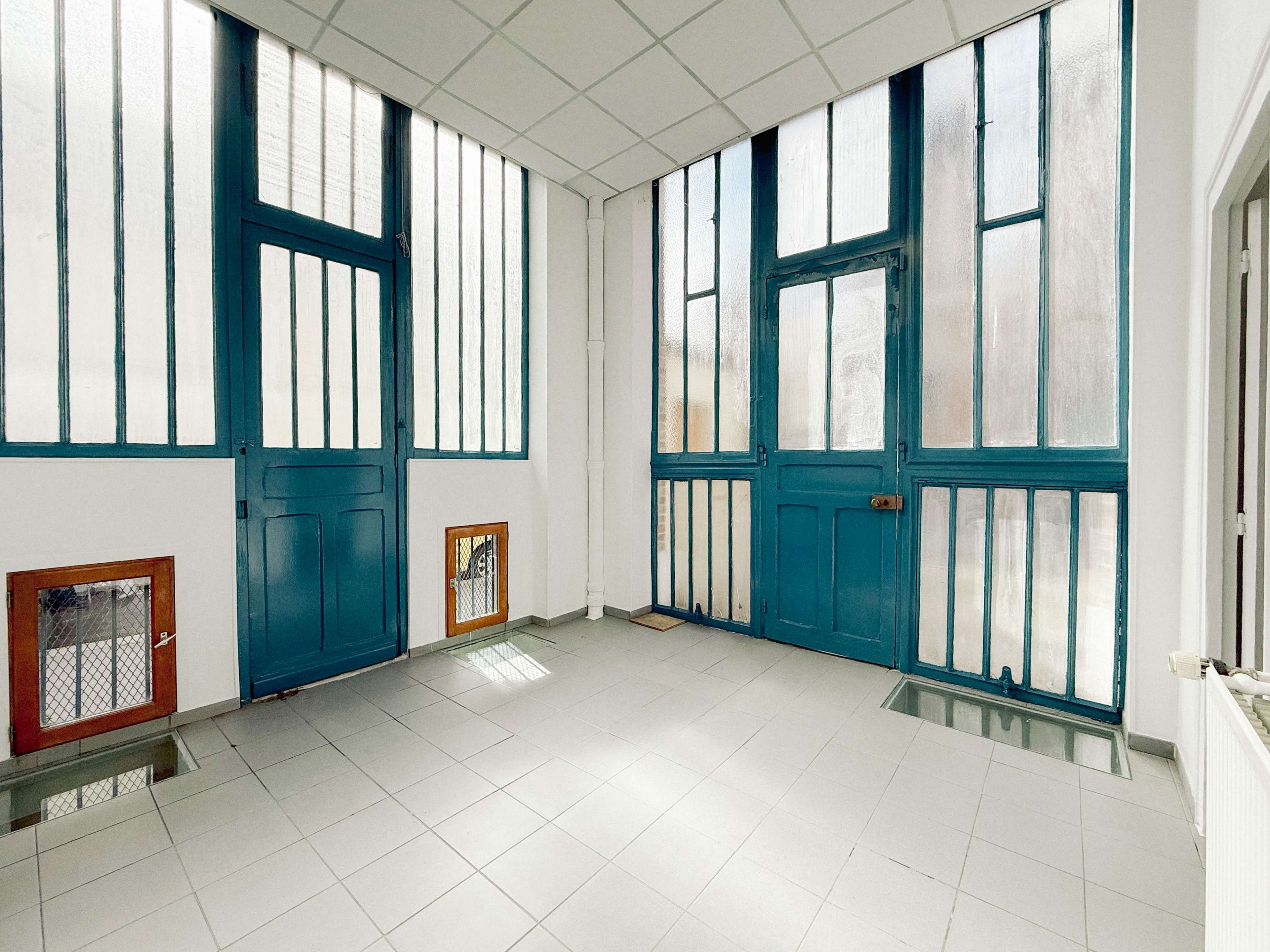 The image shows a bright, open room with large windows and teal doors on two sides, featuring a tiled floor and minimal furnishings.