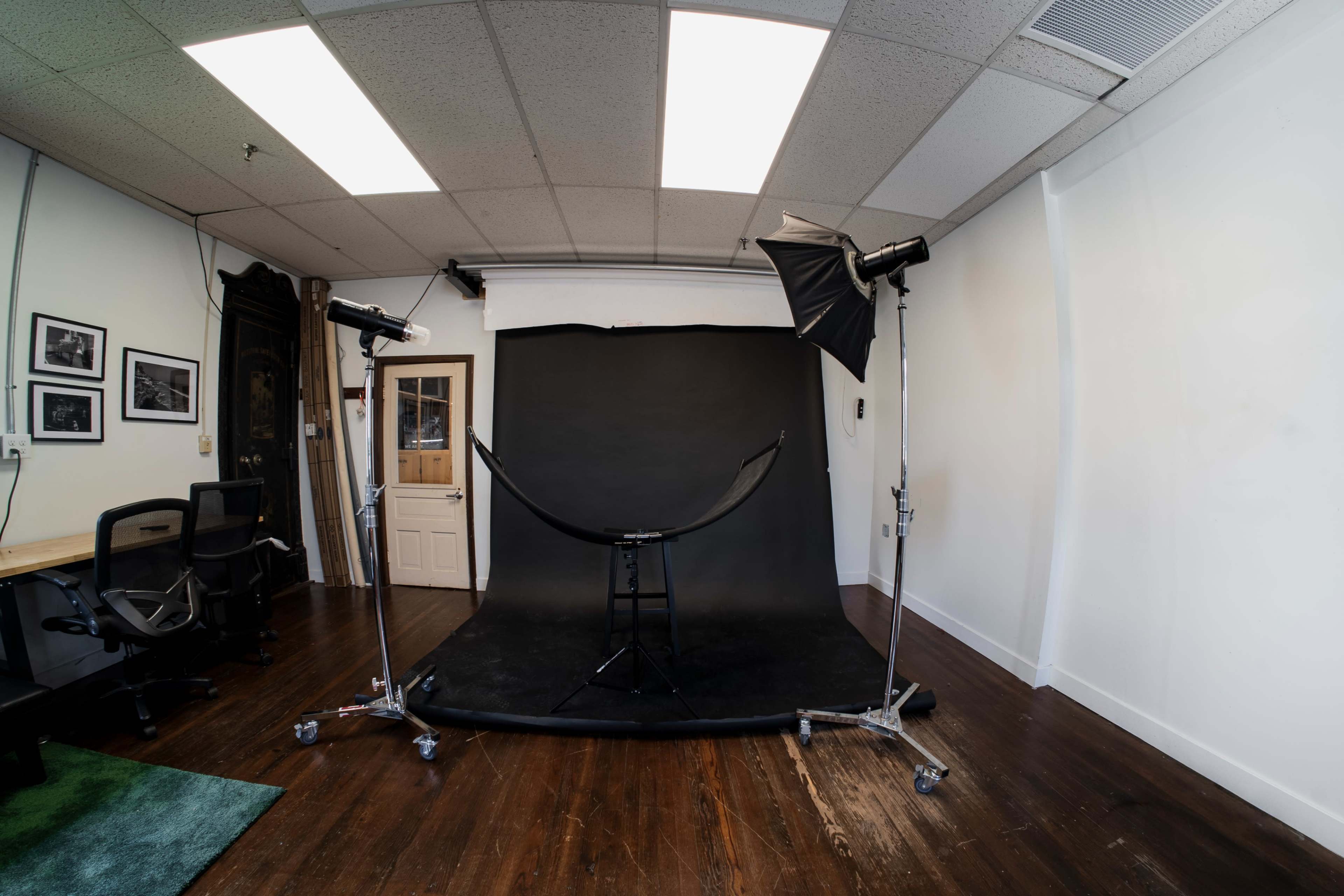 The image shows a photography studio with a black backdrop, two studio lights on stands, a stool in front of the backdrop, and a door leading to another room.