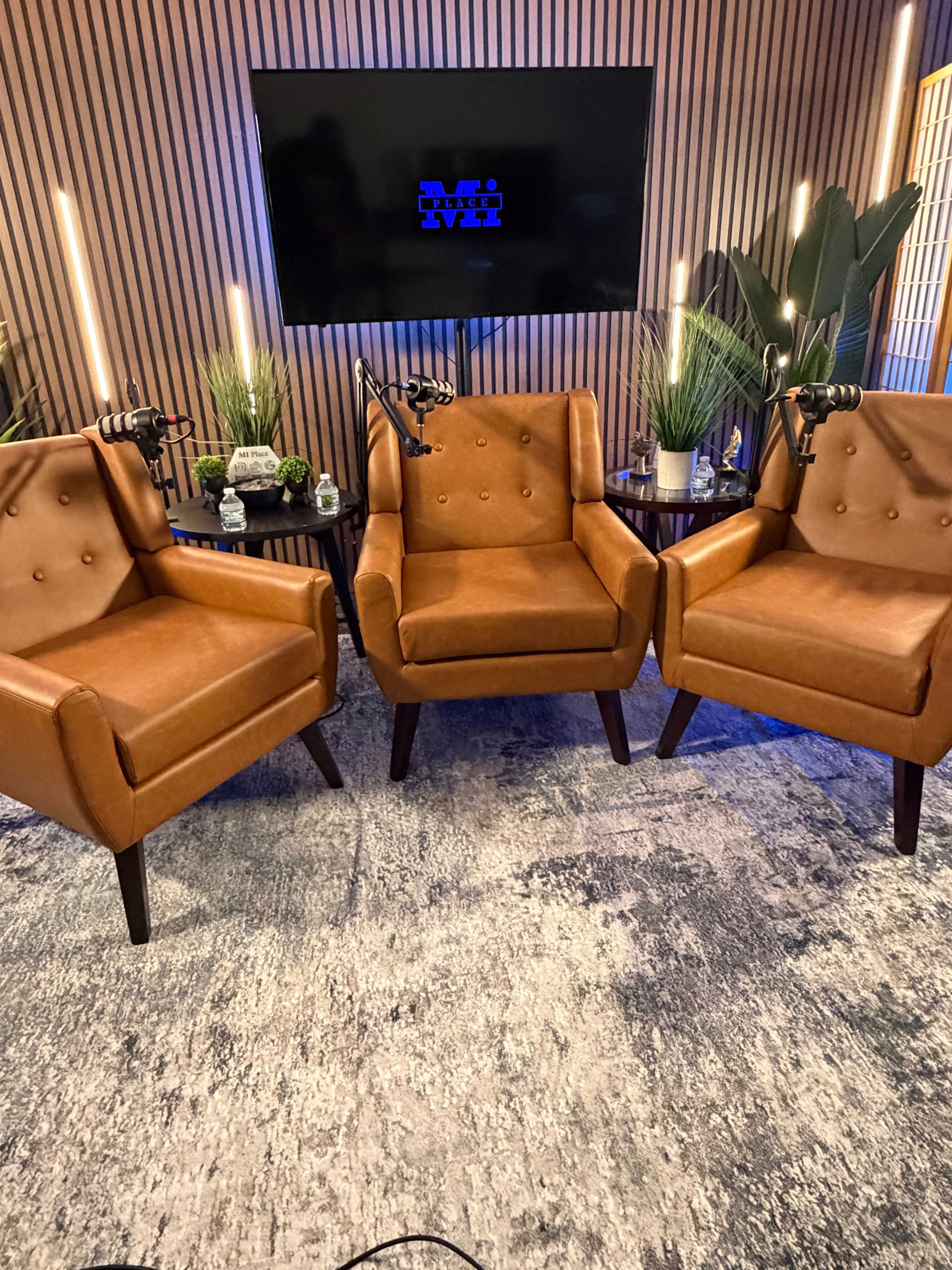 The image shows three brown leather chairs arranged in a studio setting with plants and a television screen in the background.