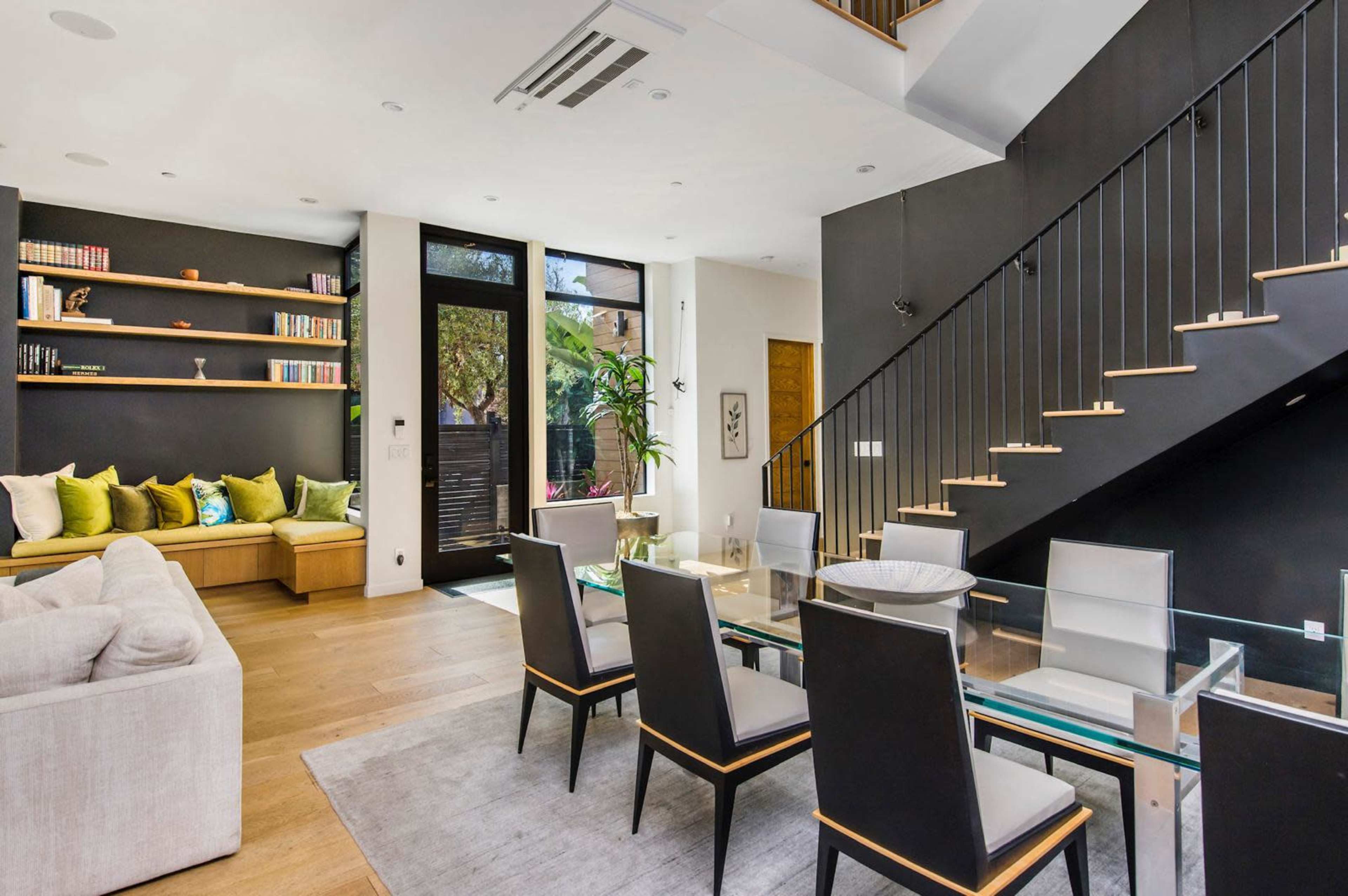 The image shows a modern living and dining area featuring a black accent wall, a glass dining table with black chairs, and a cozy seating nook with colorful cushions.