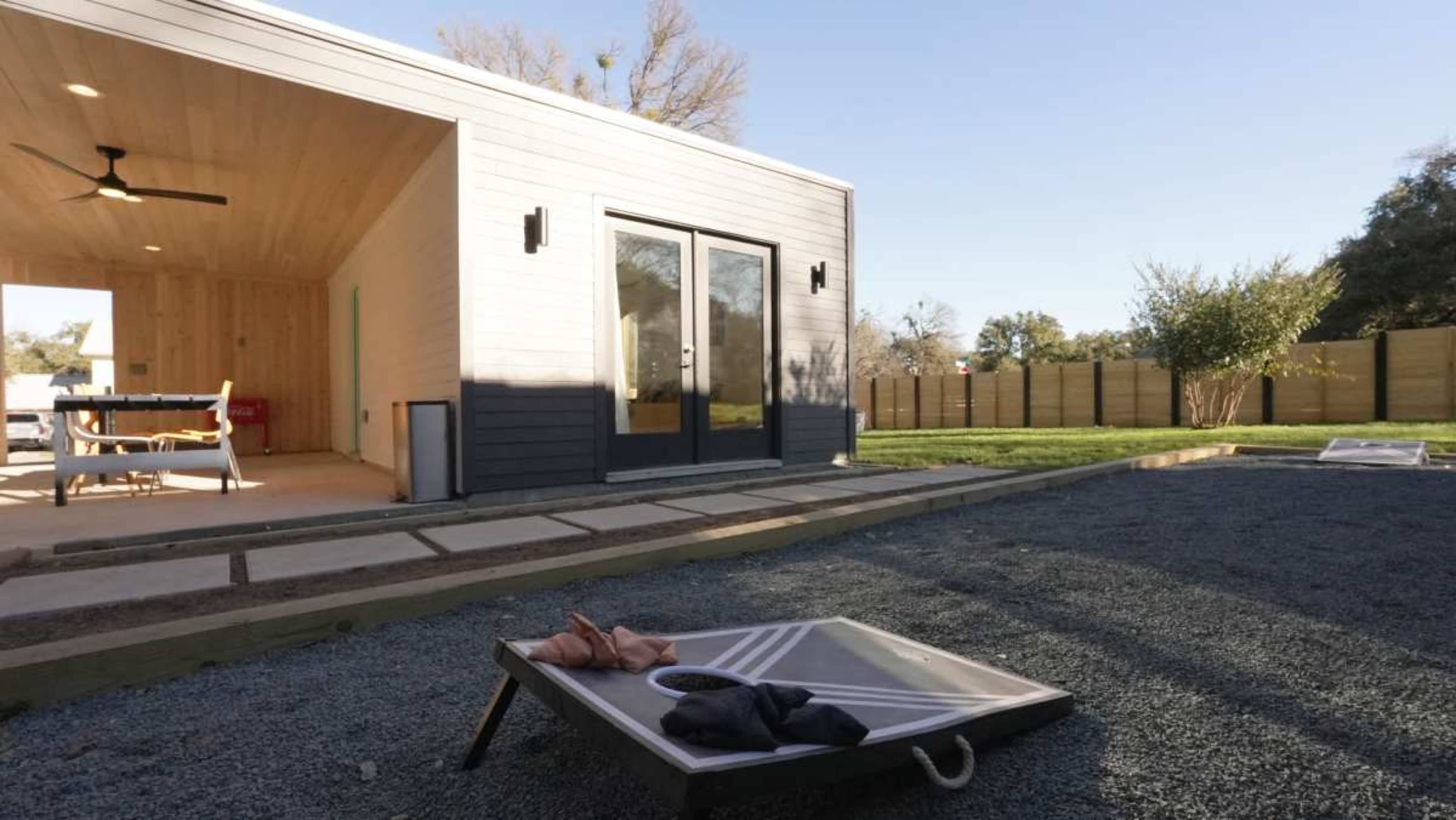 A modern backyard features a cornhole game setup on gravel, with a sleek house in the background and a spacious patio area.
