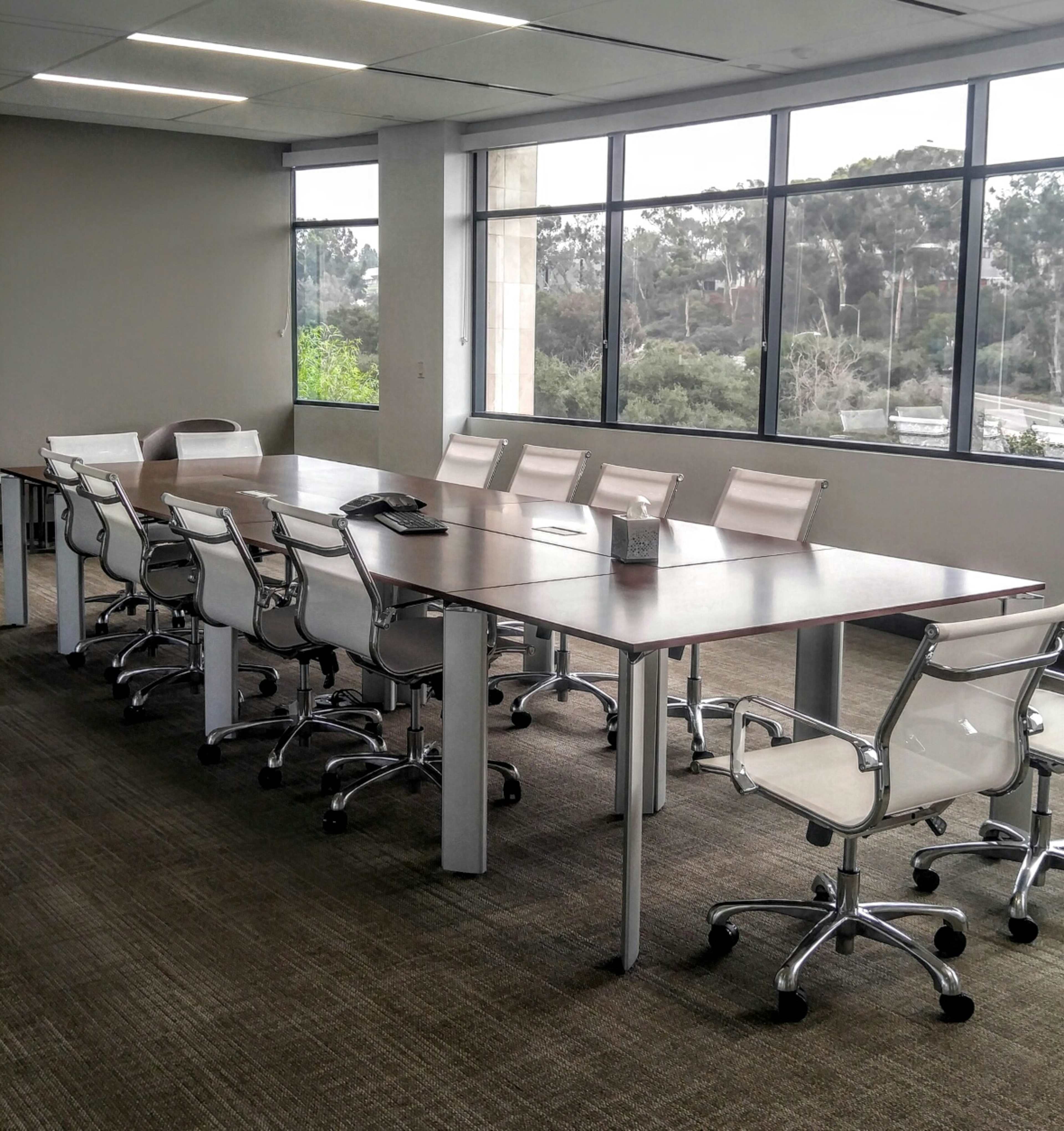 A large conference room features a long wooden table surrounded by several white ergonomic chairs, with large windows providing a view of greenery outside.