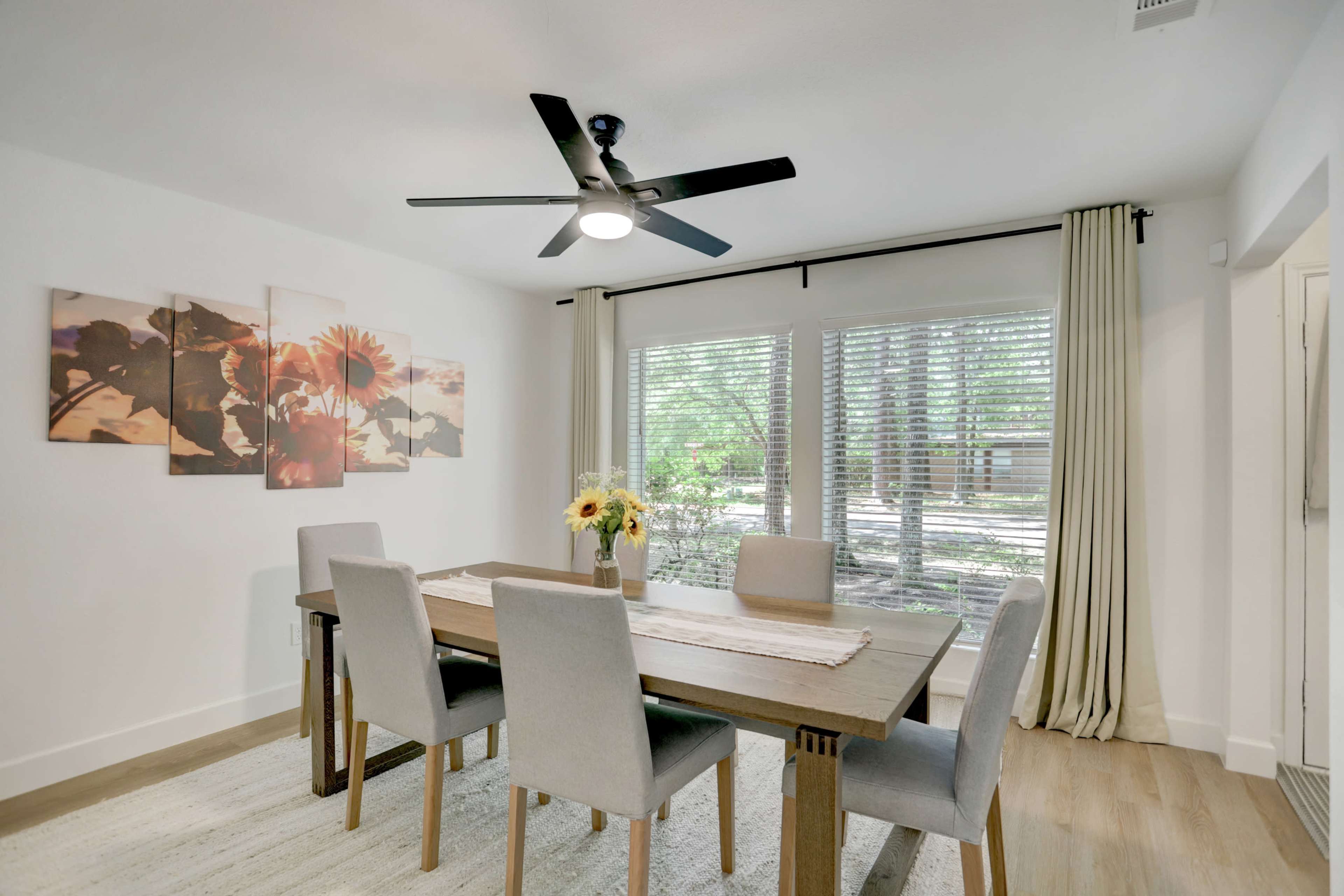 A dining room features a wooden table surrounded by four grey chairs, with a bouquet of sunflowers on the table and large windows allowing natural light to enter.