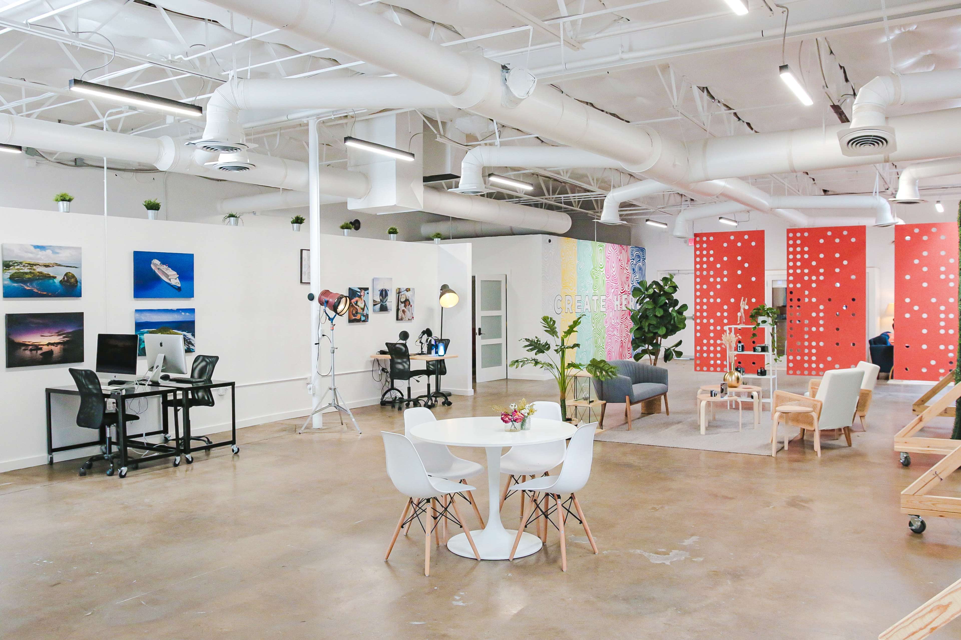A spacious, modern office environment with distinct work areas, including a meeting table, a desk with computers, and decorative walls featuring artwork.