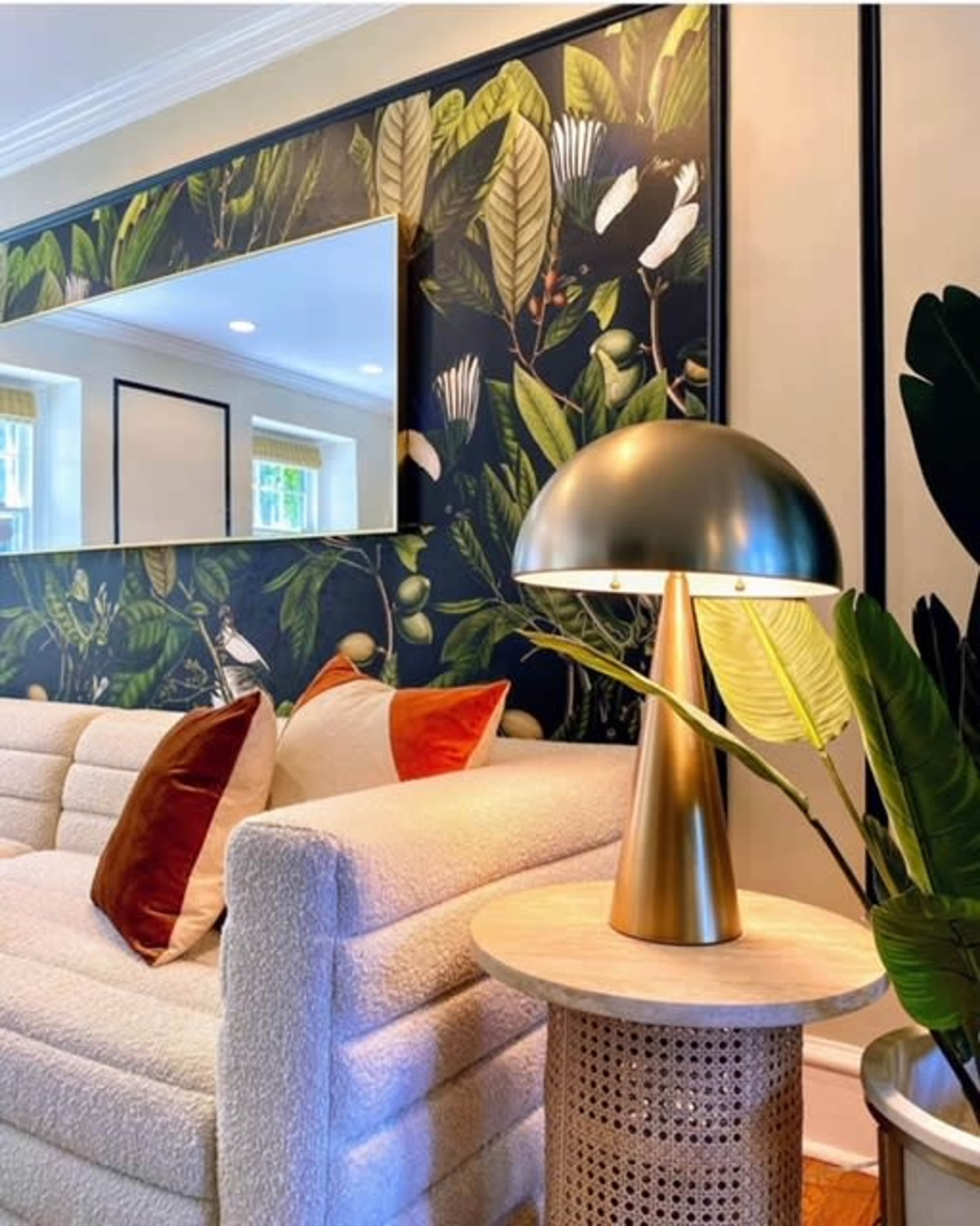 A cozy living room features a white sofa with colorful throw pillows, a modern lampshade, and a decorative plant, set against a vibrant botanical wallpaper.