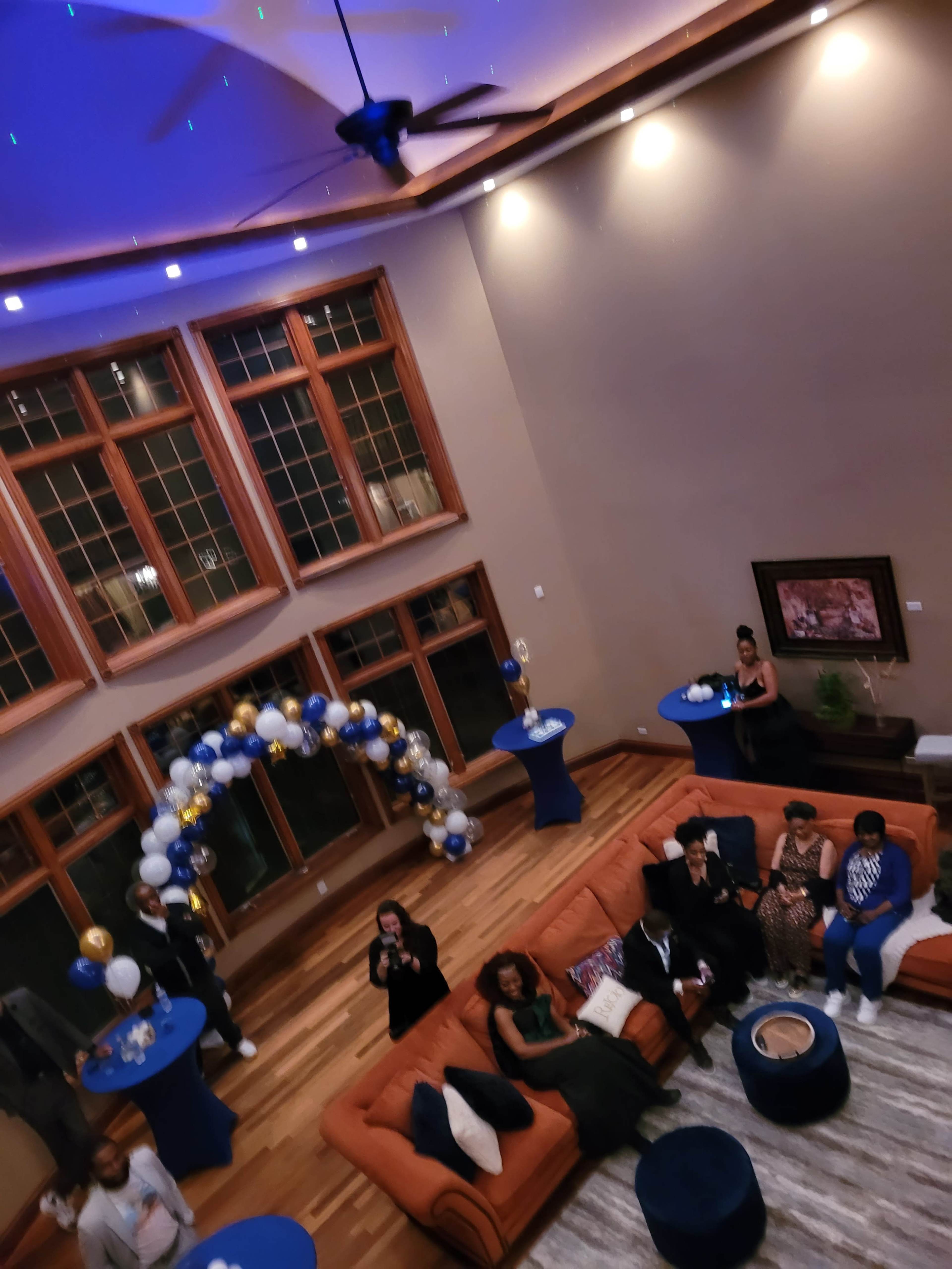 An elegantly decorated indoor space features a large sofa, multiple cocktail tables, and an arch of balloons, with guests mingling under dim lighting.