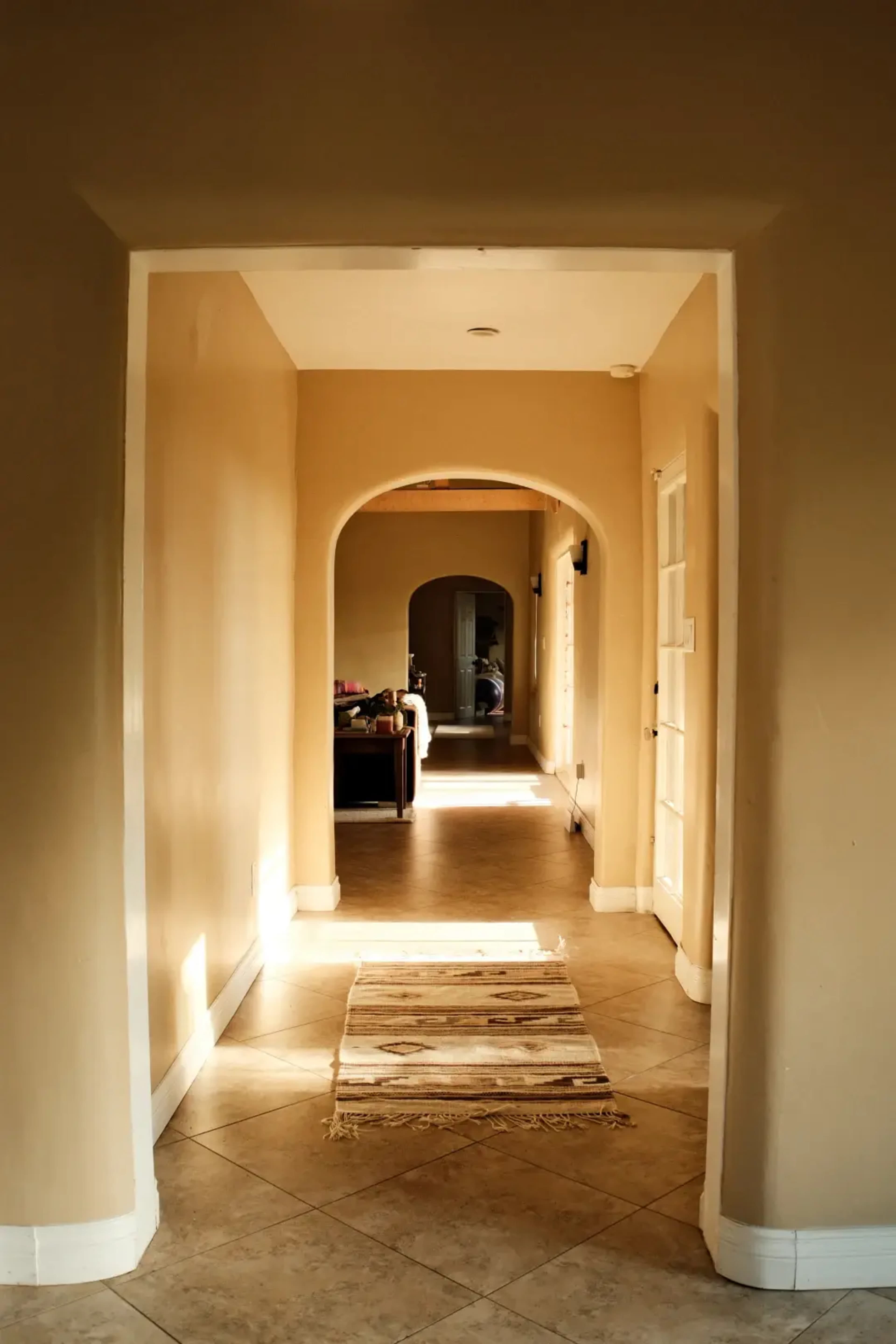 The image shows a long hallway with a patterned rug on the tiled floor, leading to an arched opening and a glimpse of a living area at the end.