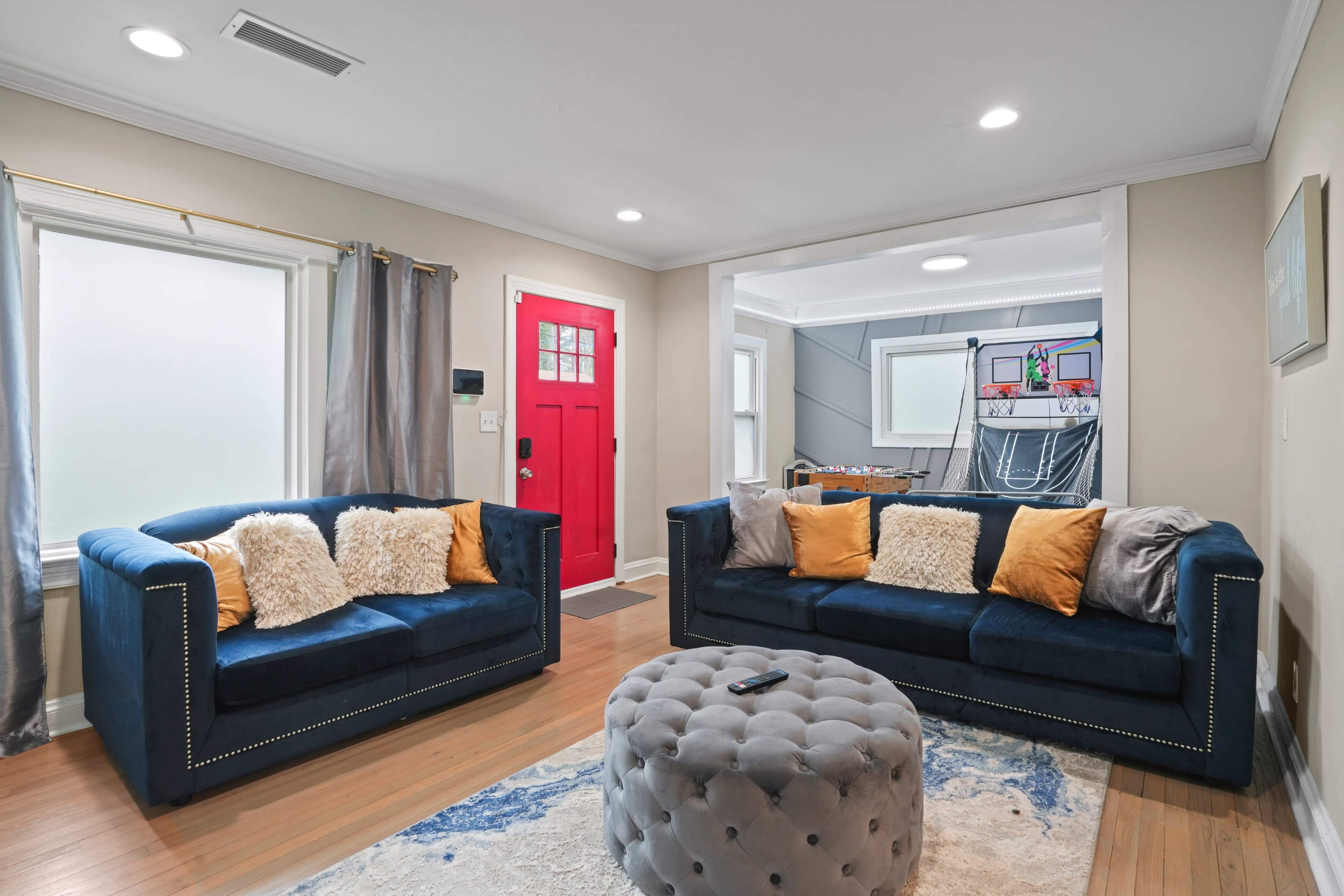 The living room features two navy blue sofas with decorative pillows, a round ottoman in the center, and a bright red door in the background.