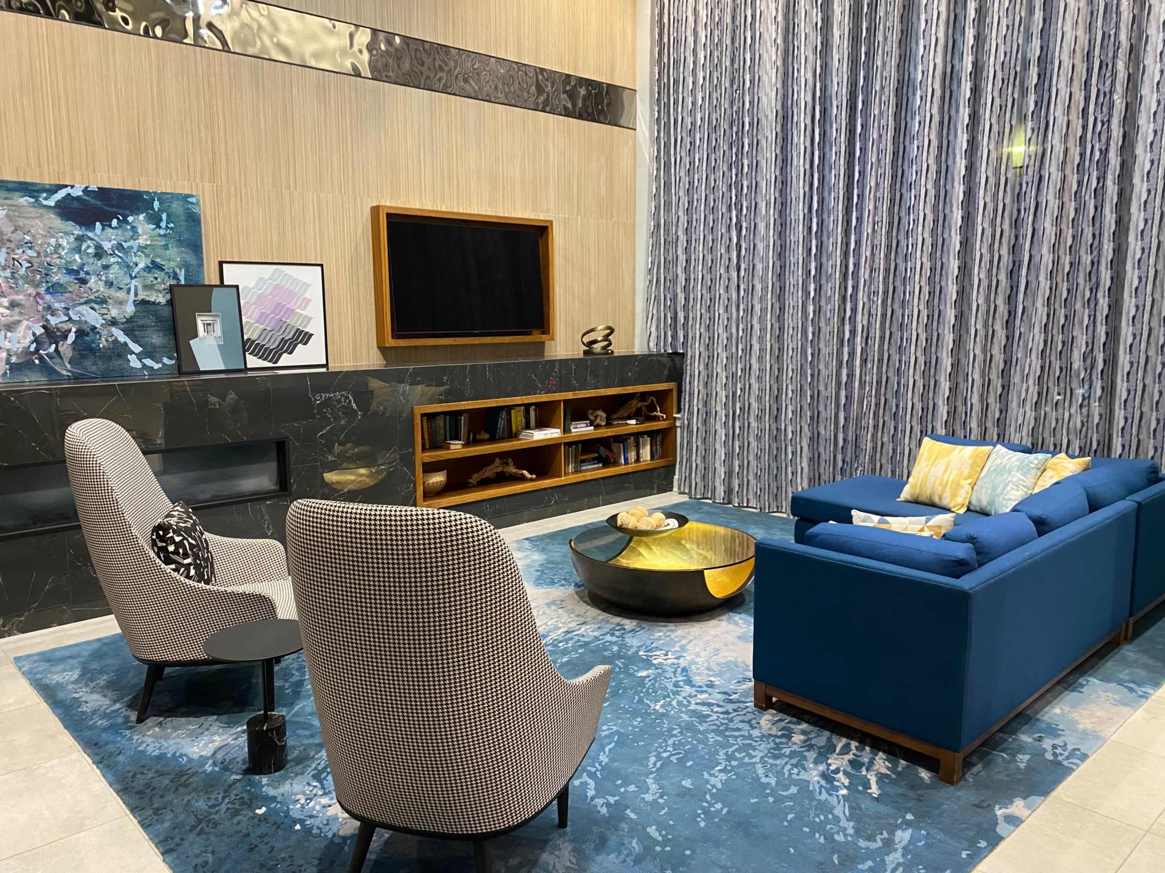 A modern living room with two patterned chairs, a blue couch, a round coffee table, and a wall-mounted television, set against a backdrop of vertical curtains and a dark marble feature wall.
