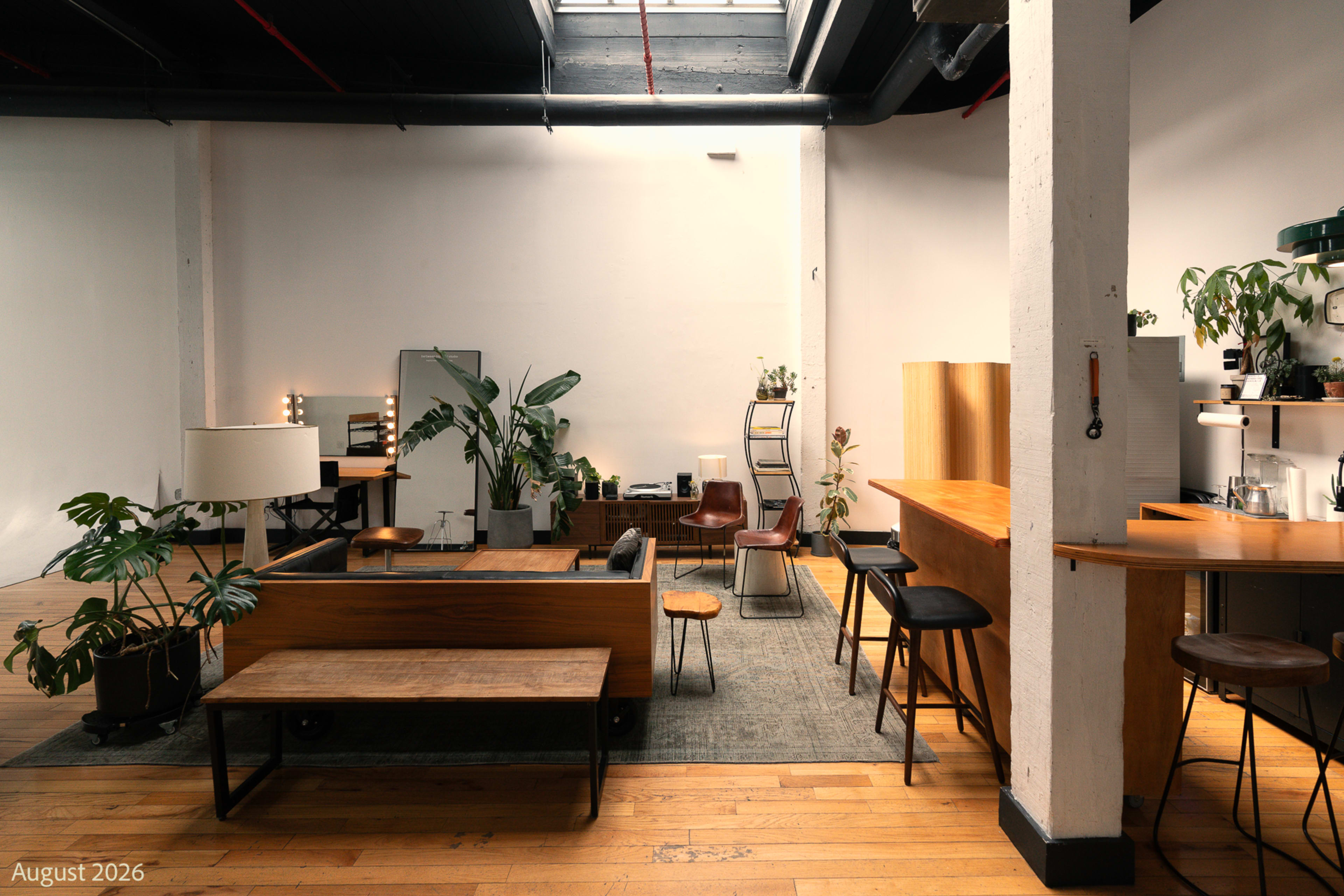 The image shows a spacious, modern interior with wooden flooring, a mix of seating options, houseplants, and various decorative elements.
