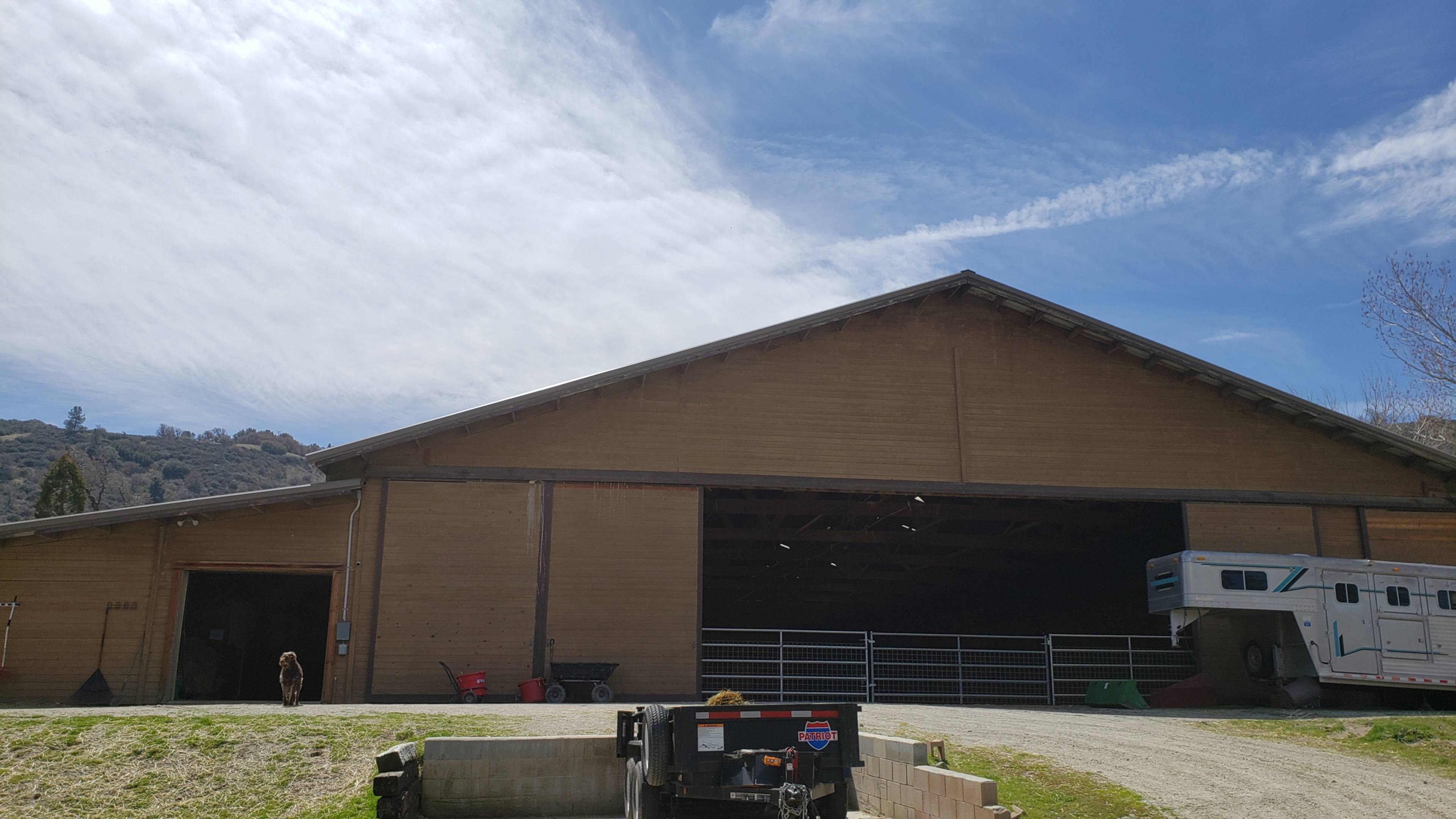 A large wooden barn with an open front, located on a gravel area, alongside a horse trailer and various tools.